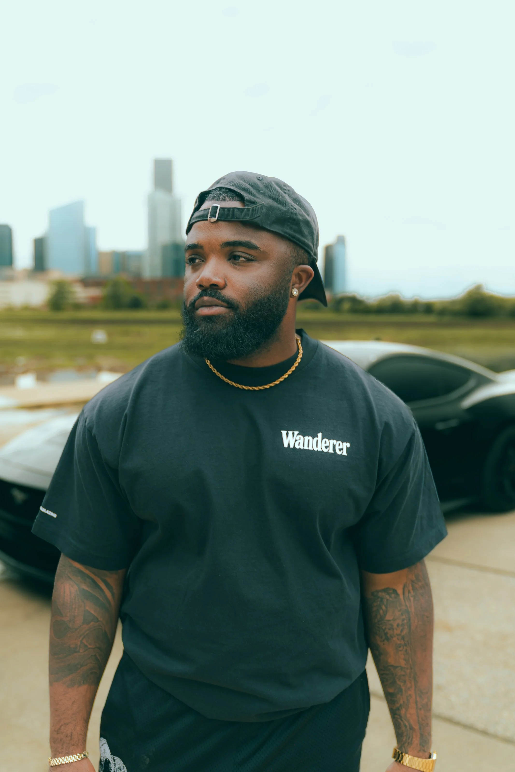 A man with a beard wearing a black t-shirt with the word 'Wanderer' on it, a backwards black cap, gold chain, gold watch, and earrings, standing outdoors with a city skyline in the background.
