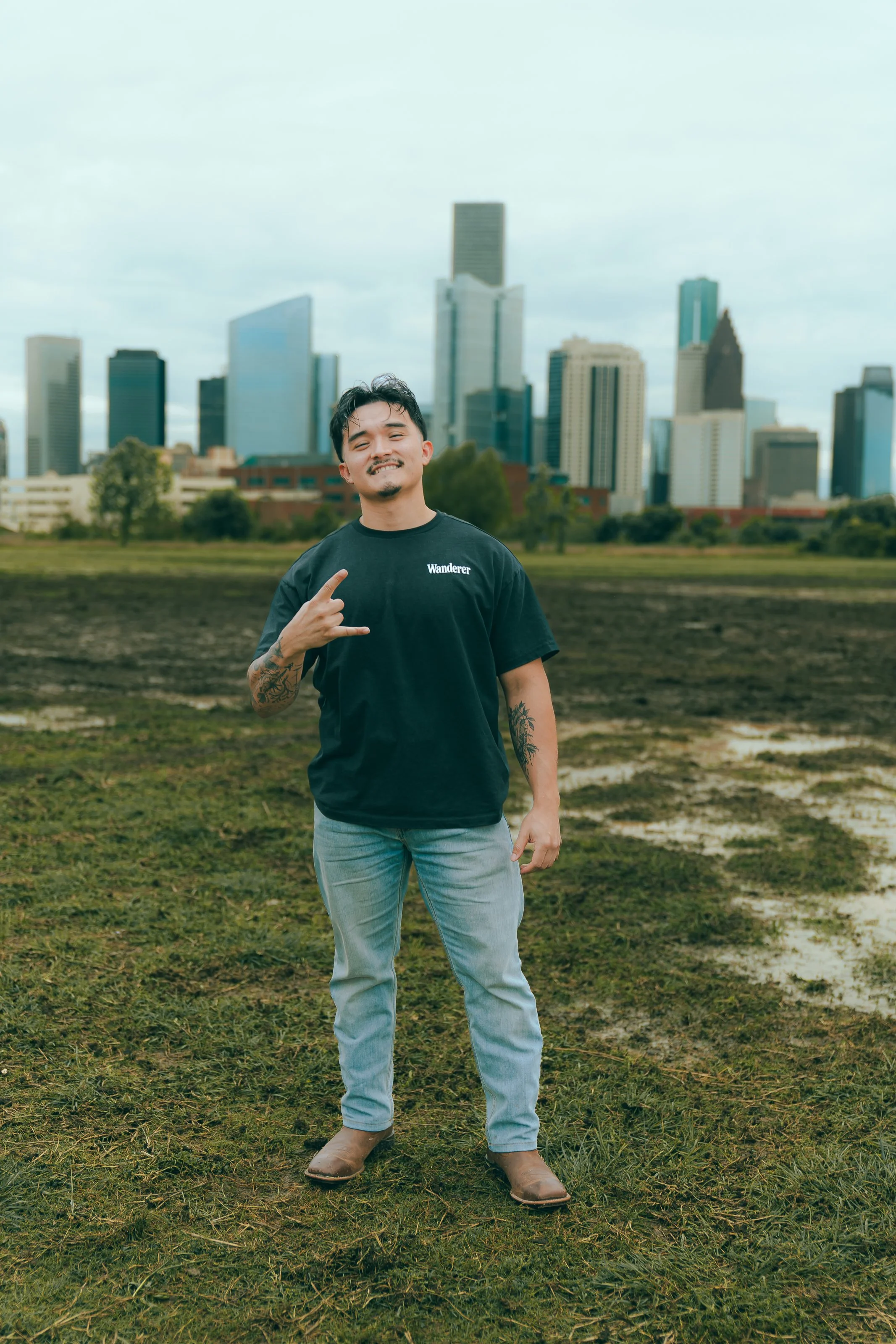 A man with tattoos on his arms, wearing a black t-shirt with 'Wanderer' written on it, light blue jeans, and brown boots, standing in an open grassy area with a city skyline of tall buildings in the background on a cloudy day.