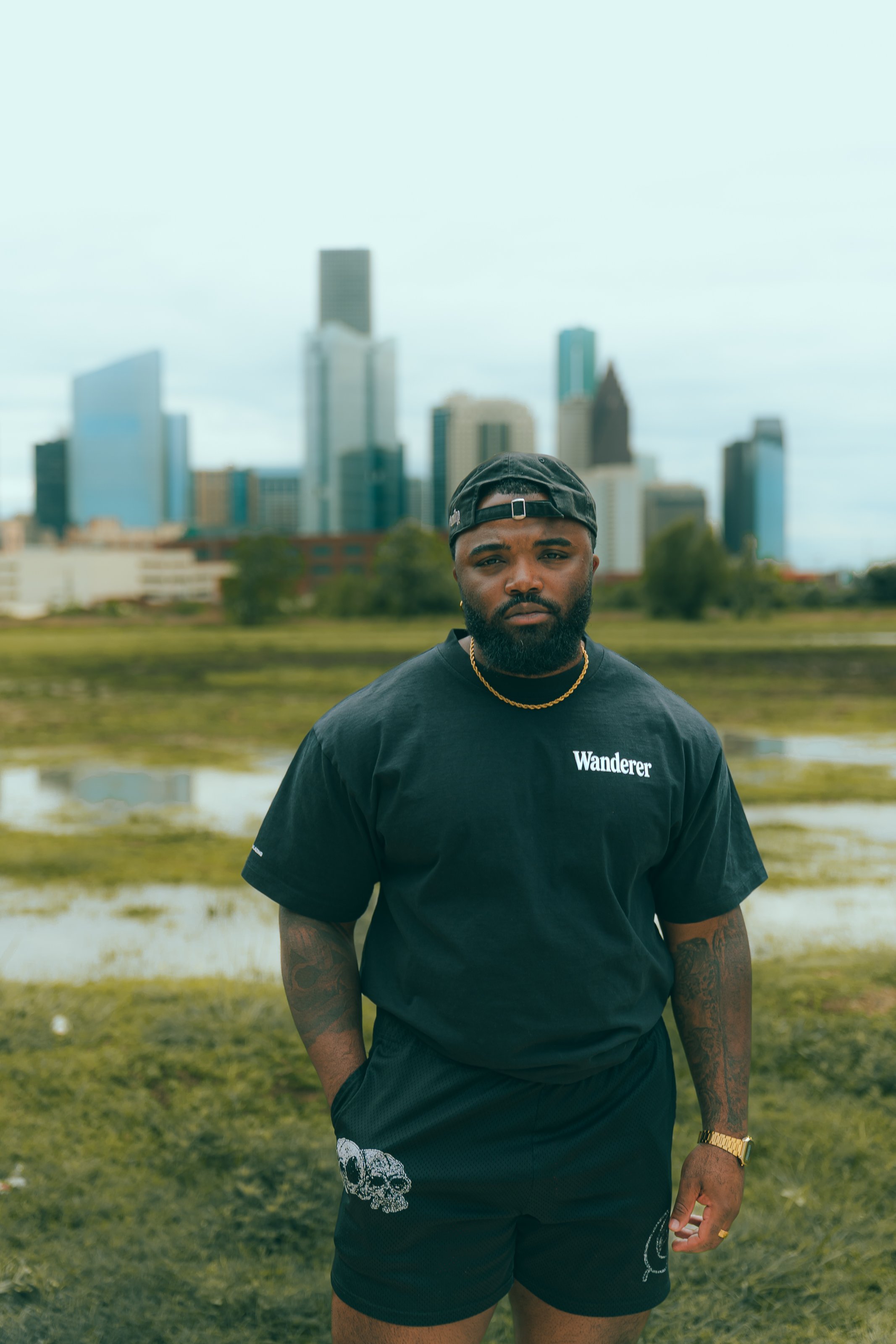 A man standing outdoors in front of a city skyline during daytime. He has a beard, tattoos on his arms, is wearing a black baseball cap backwards, a gold chain, a black T-shirt that says 'Wanderer', and black shorts with a skull design.