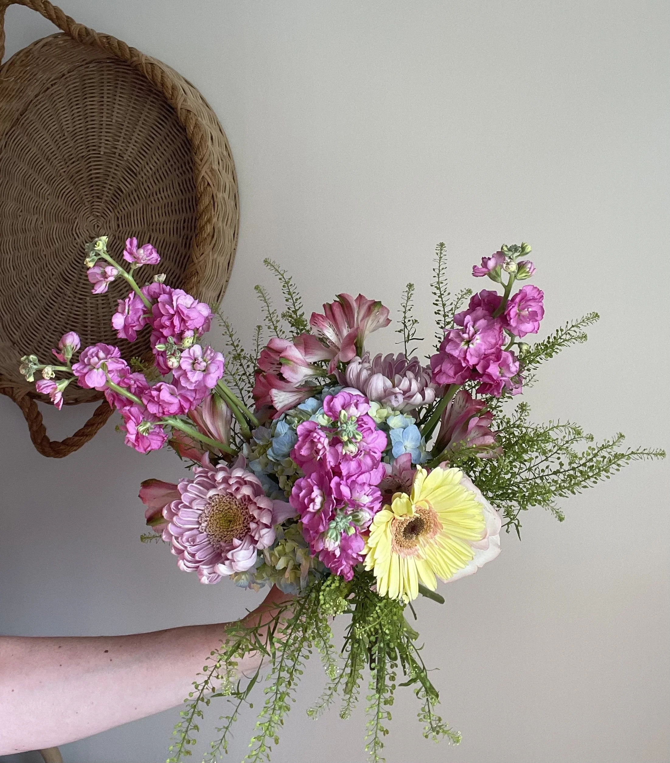 A person holding a bouquet of pink, yellow, and purple flowers with green foliage, against a white wall and a woven basket in the background.