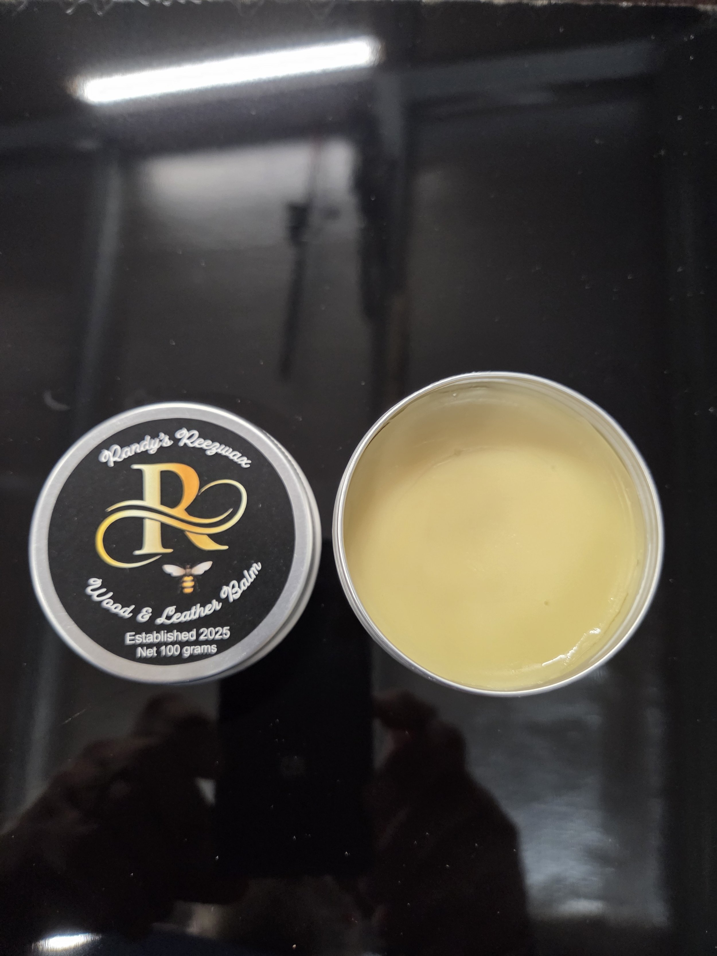 Open tin of balm next to its lid on a black surface.