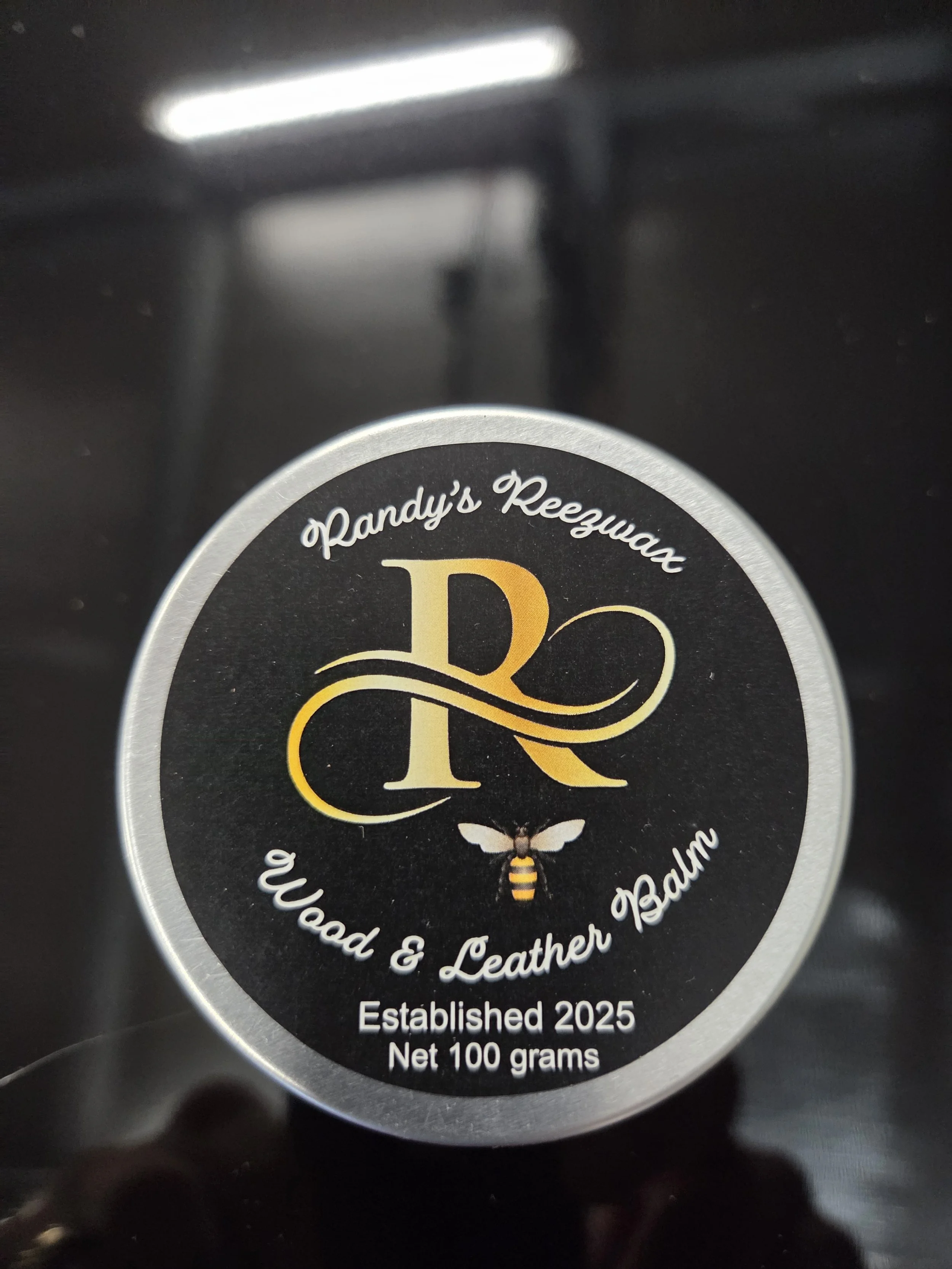Close-up of a circular product label with black background and silver border, featuring gold and white text, a large decorative 'R' with swirls, a bee illustration, and details indicating it is from Randy's Reezwax, a wood and leather balm established in 2025, weighing 100 grams.