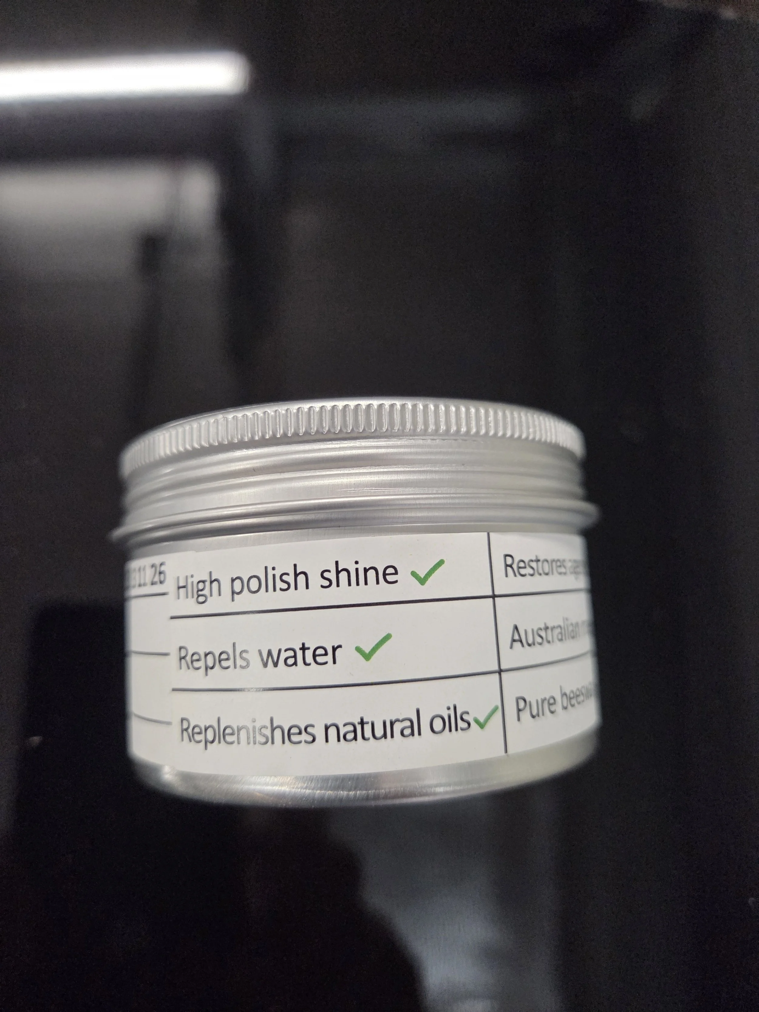 Close-up of a small white container with a silver screw-on lid, labeled with benefits of natural skincare ingredients such as high polish shine, water repellence, and natural oils, placed on a dark surface.