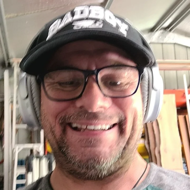 A man with glasses, a beard, and a smile, wearing a black baseball cap and large headphones, taking a selfie in a workshop environment.