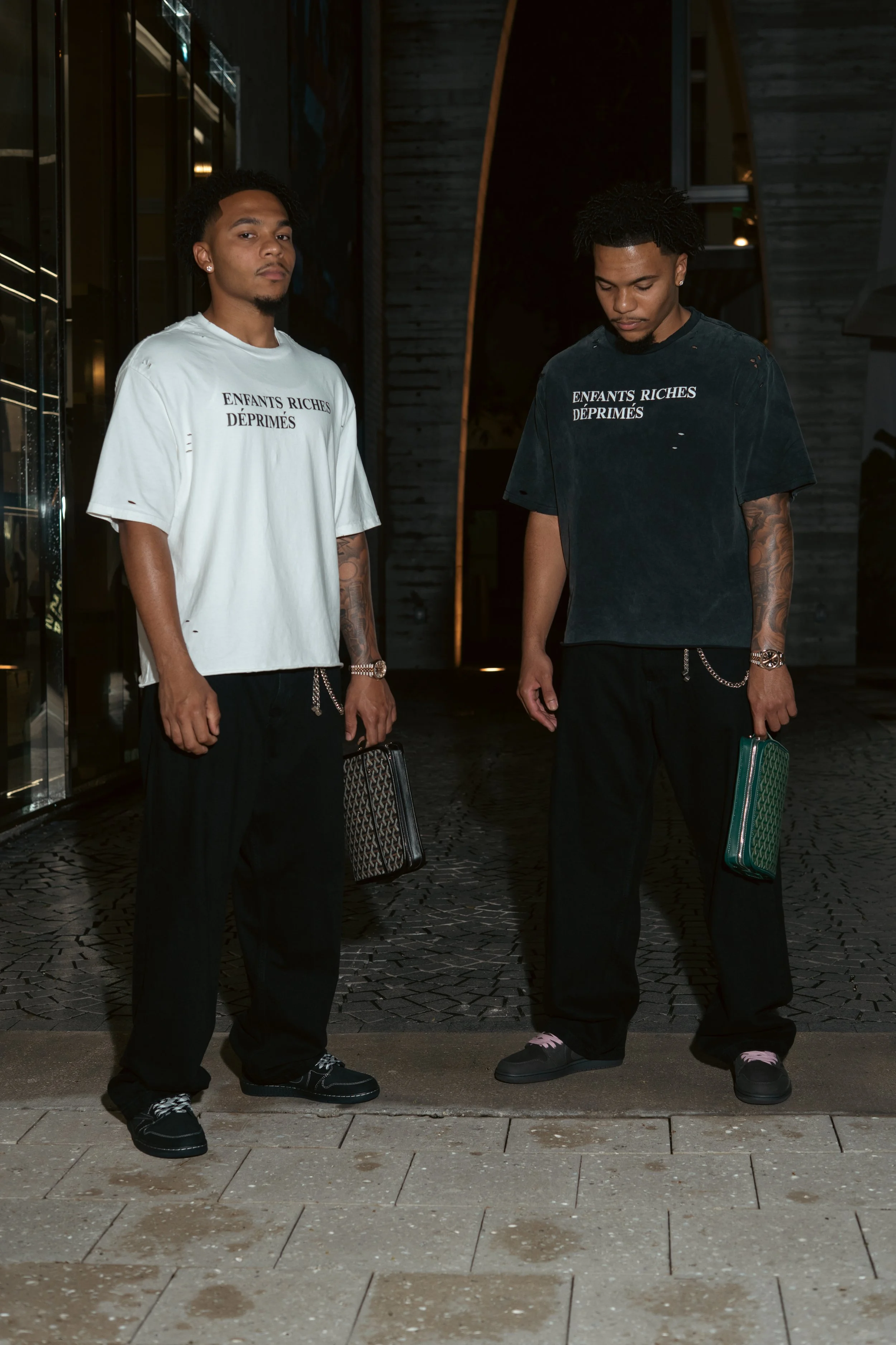 Two young men standing outdoors at night, wearing oversized graphic t-shirts with the phrase 'ENFANTS RICHES DÉPRIMÉS,' black pants, and holding designer bags. One has tattoos on his arm and wears a watch, while the other has curly hair and is lookin