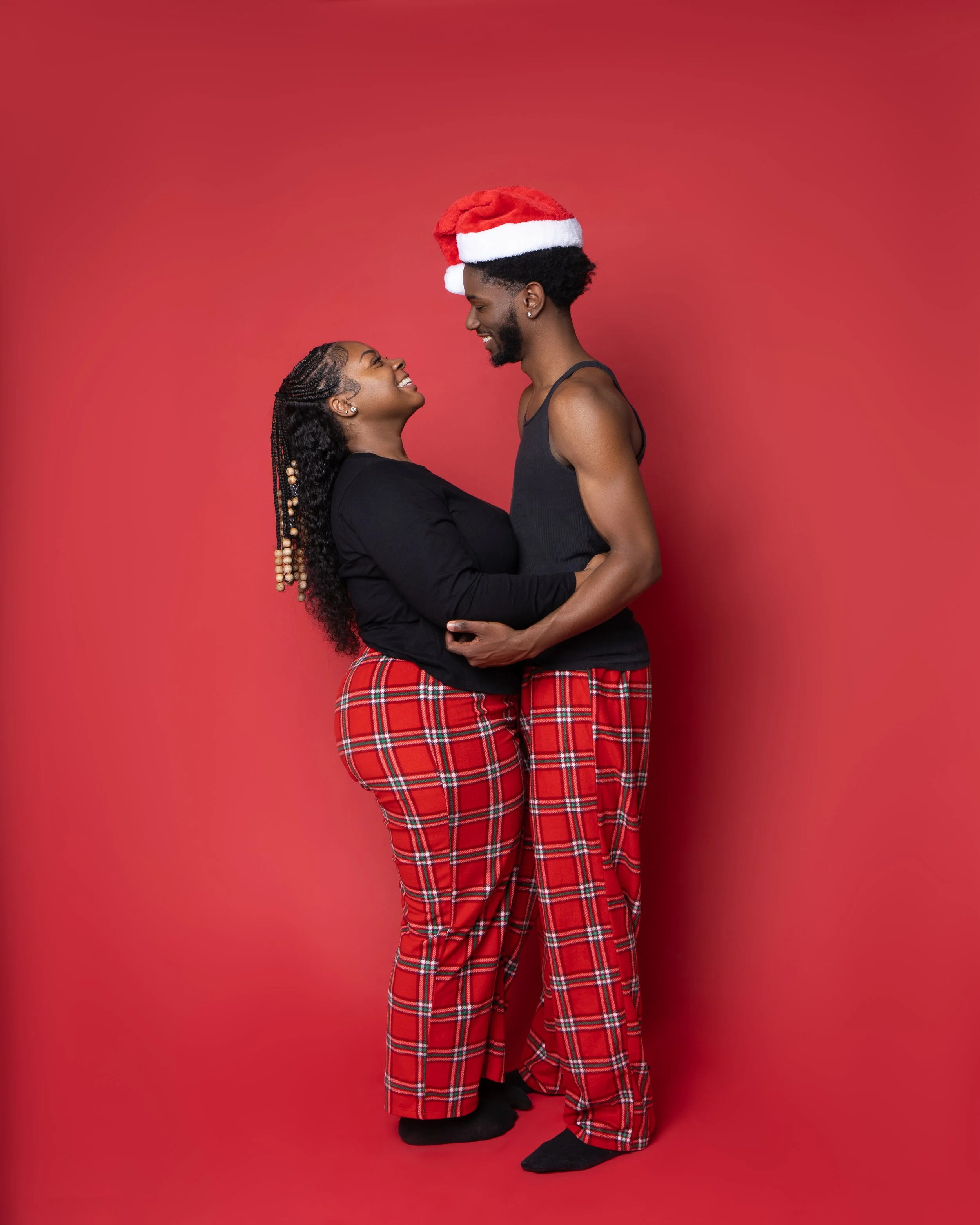 A couple in matching red plaid pajamas and Santa hat posing against a red background, smiling and embracing each other.