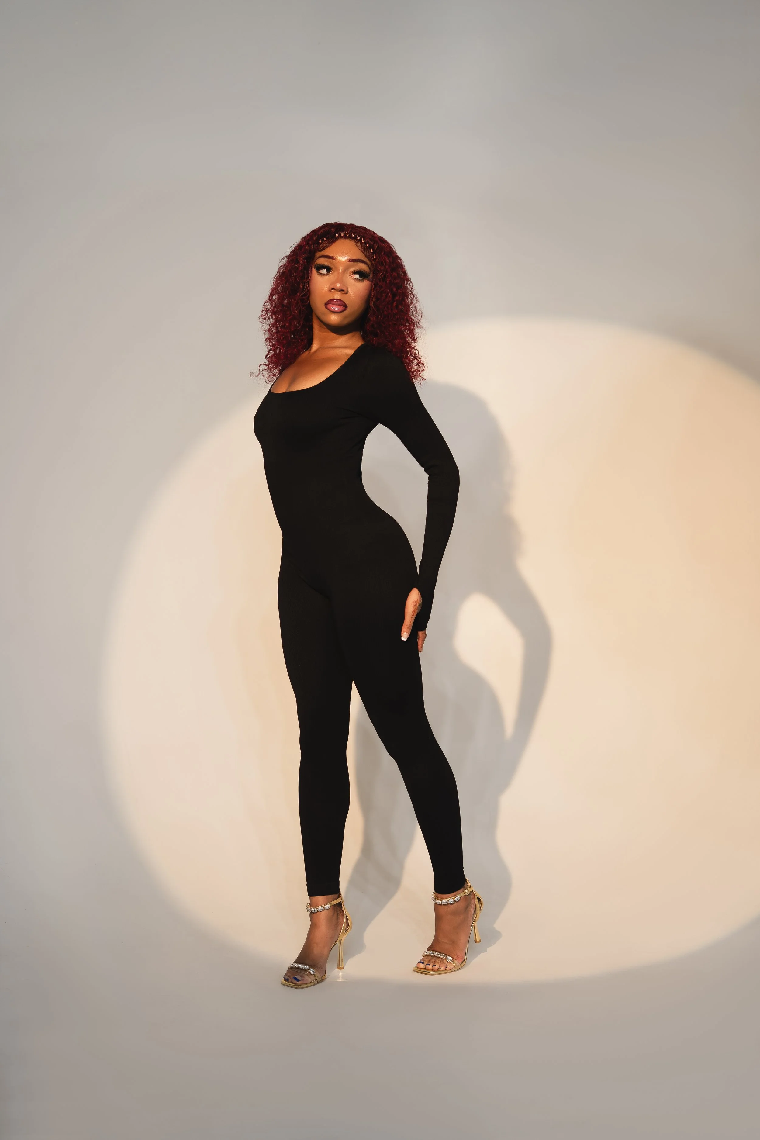 A woman with curly red hair in a black bodysuit and high heels posing against a plain background with a spotlight effect.