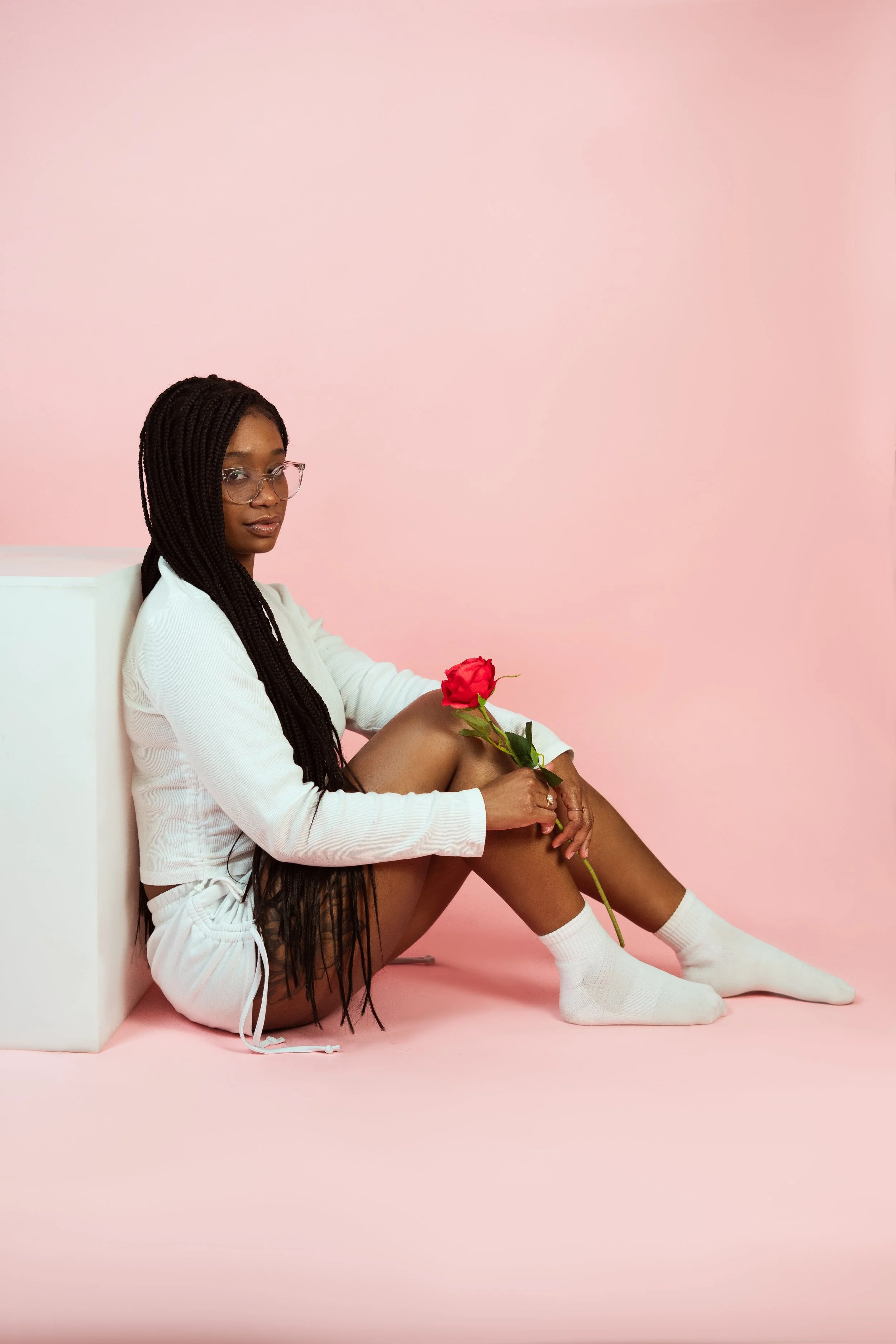 A woman with long braided hair and glasses sitting on the pink floor against a pink wall, wearing white socks, white outfit, holding a red rose.
