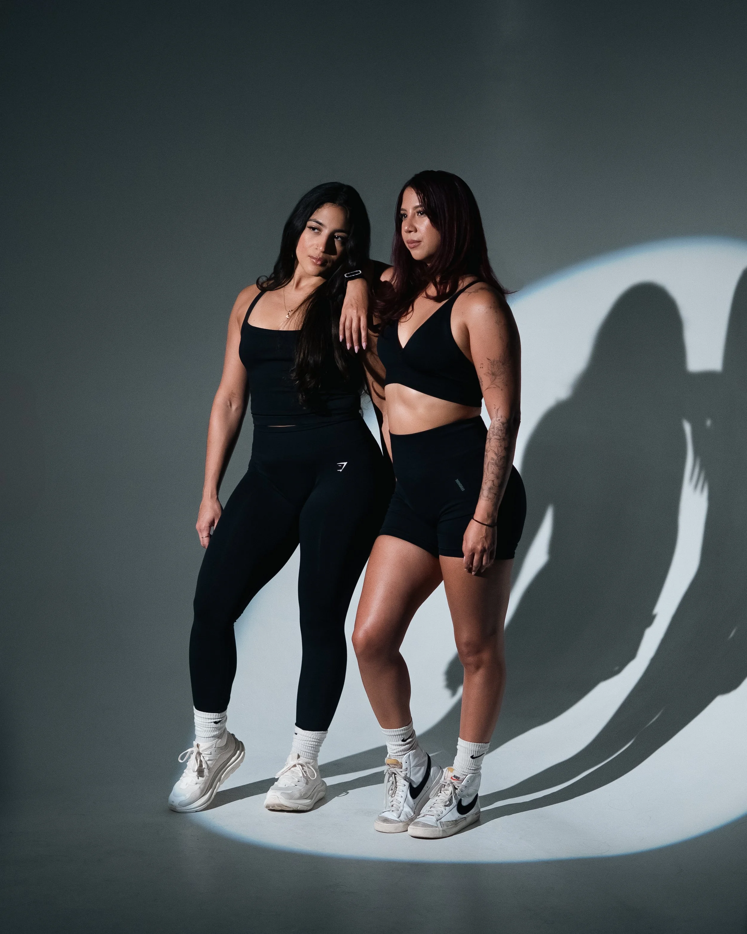 Two women standing against a dark background, cast shadows visible on the wall, dressed in black athletic wear and white sneakers.