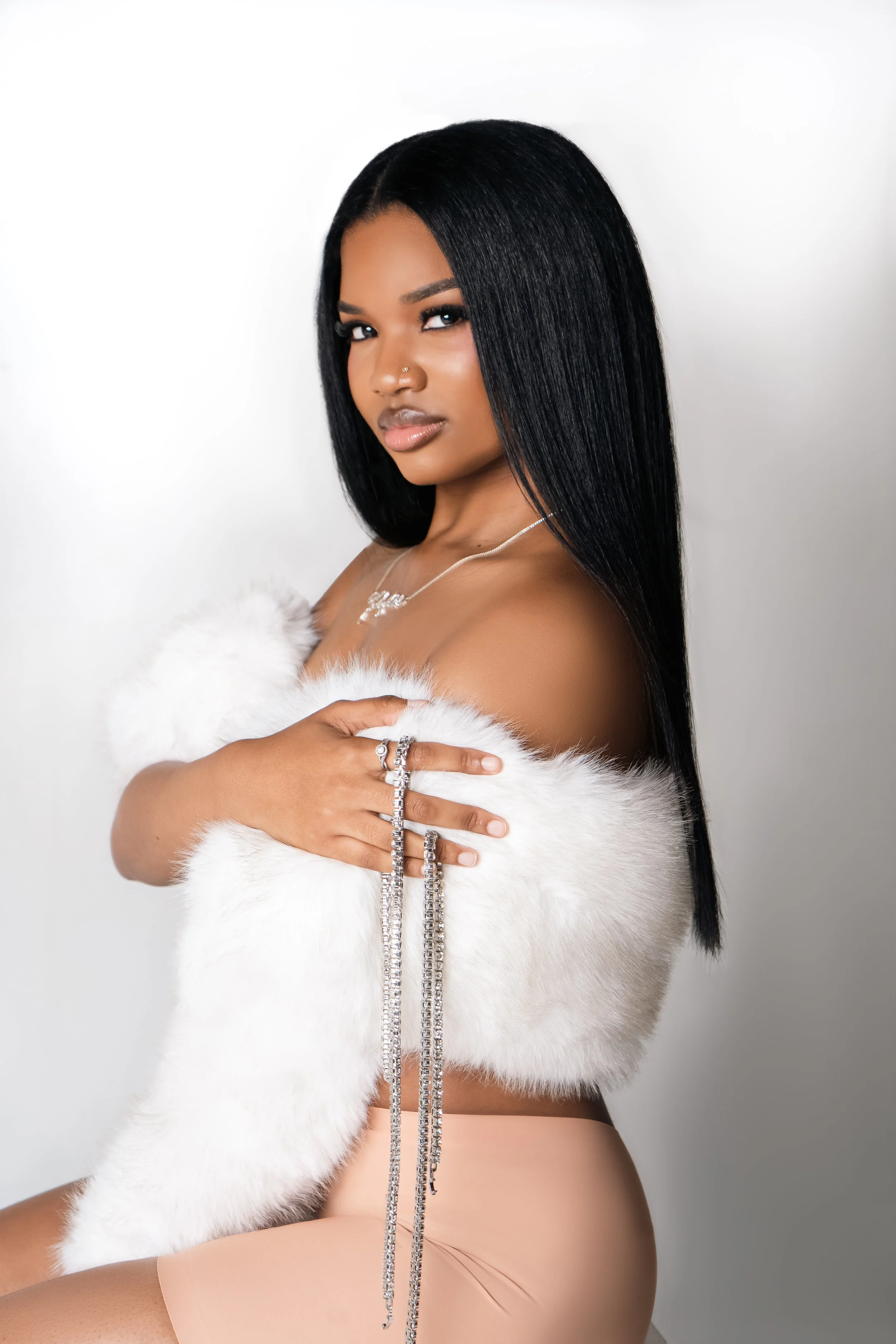 A woman with long straight black hair, wearing a white fur stole, nude-colored clothing, and silver jewelry, posing against a plain white background.