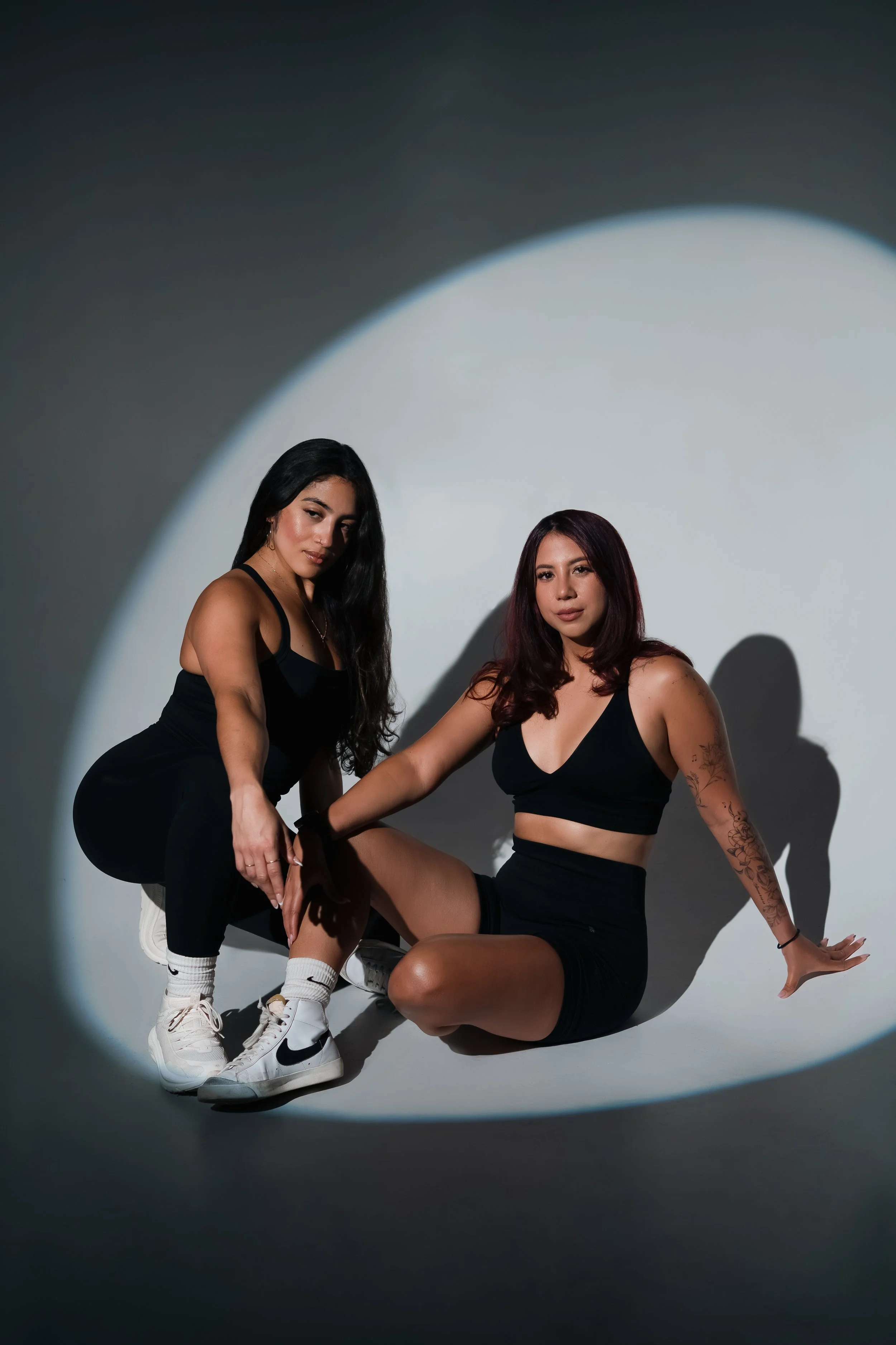 Two women in black sportswear pose on a circular spotlight against a plain backdrop.