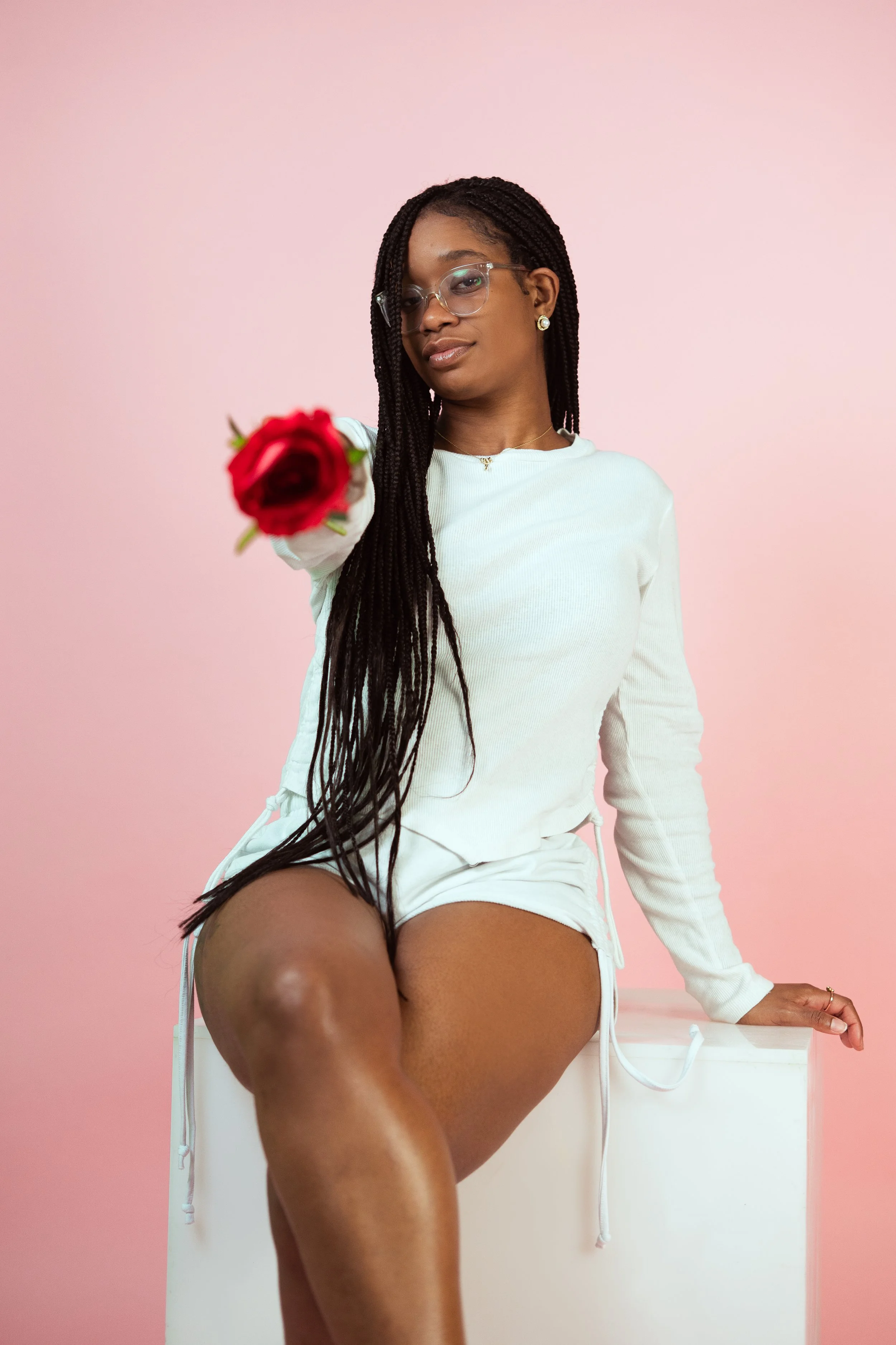 A young woman with long braided hair, wearing glasses and earrings, sitting on a white block, holding a red rose towards the camera, against a pink background.