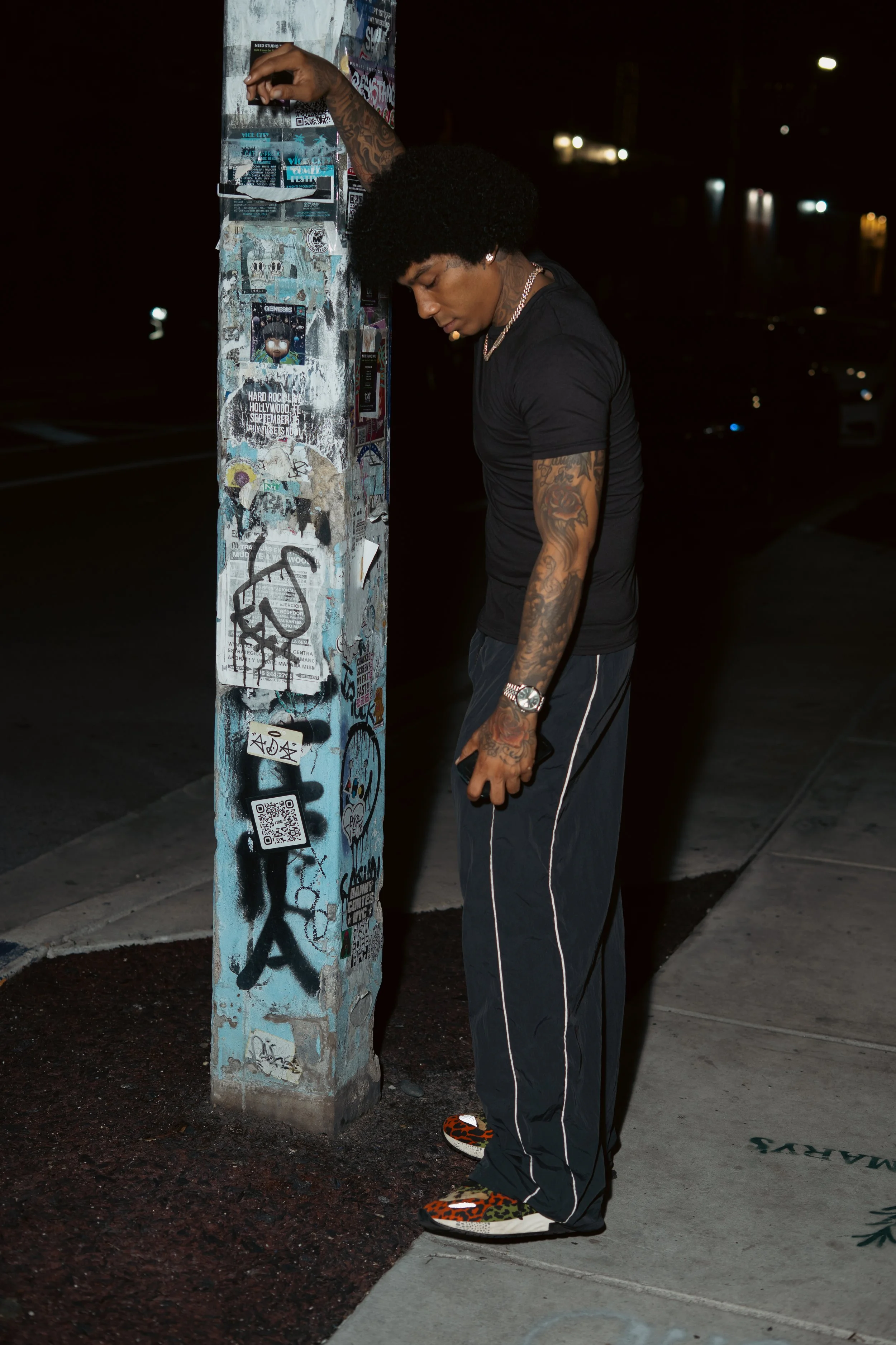 A young man with tattoos, black curly hair, dressed in black shirt and pants, wearing colorful sneakers, leaning against a graffiti-covered pole on a city sidewalk at night.