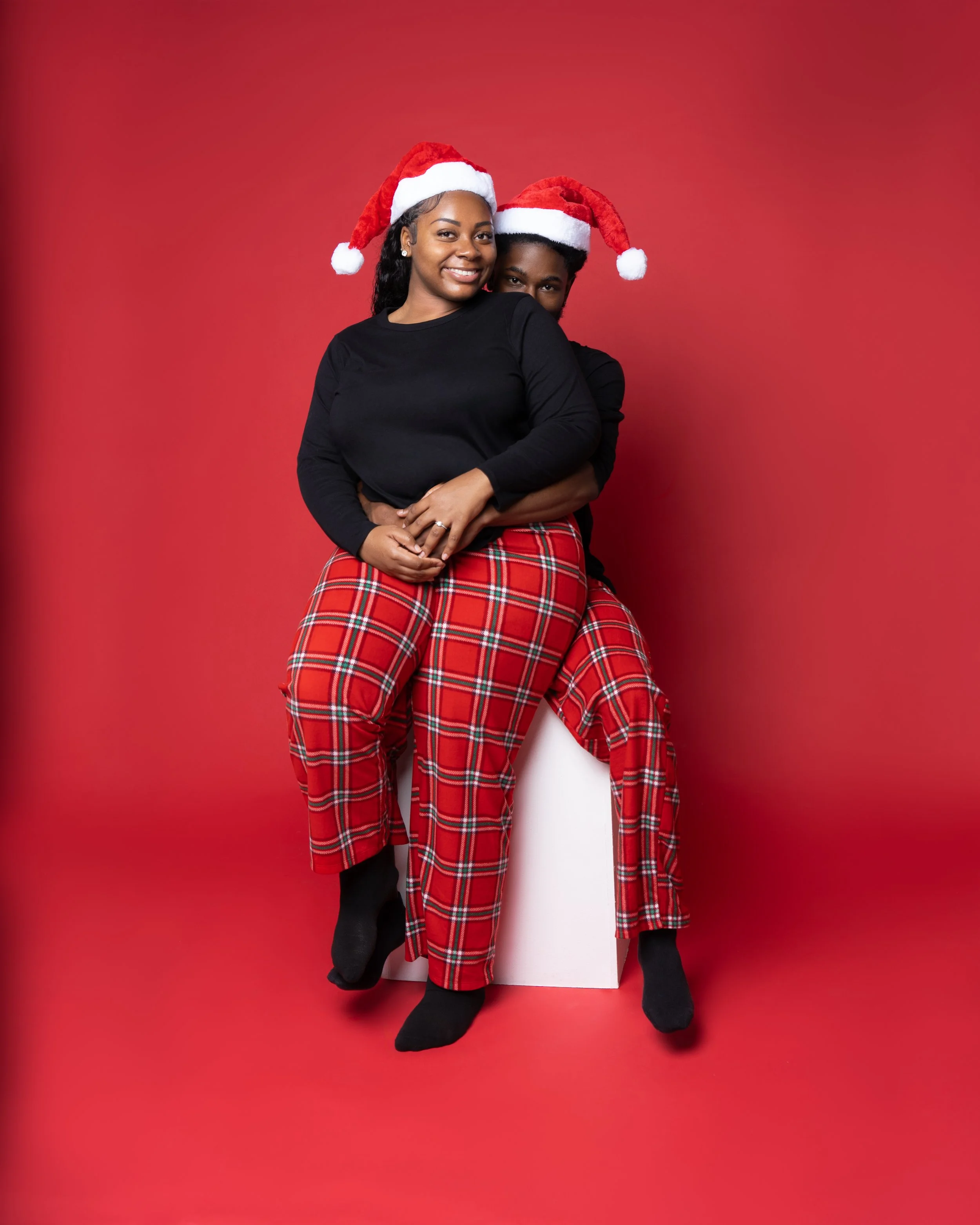 A smiling woman and man wearing Santa hats, sitting together against a red background. The woman is in front, wearing a black top and red plaid pants, while the man is behind her, partially hidden.