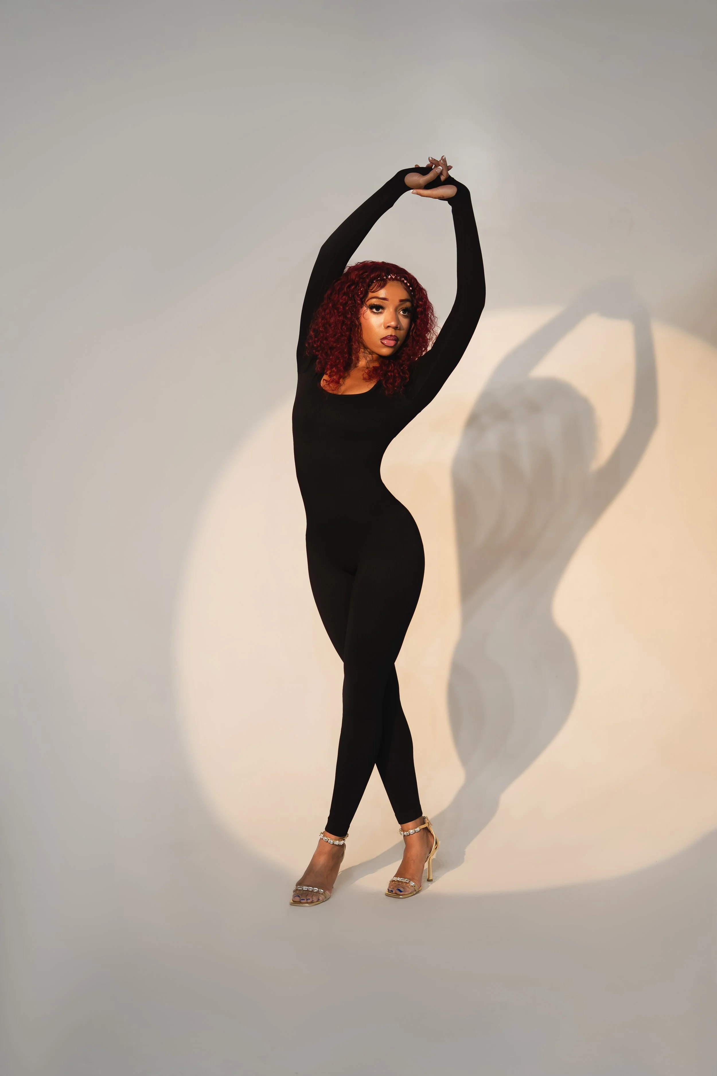 A woman in a black jumpsuit and high heels poses with her arms raised above her head, creating a shadow on the plain background.
