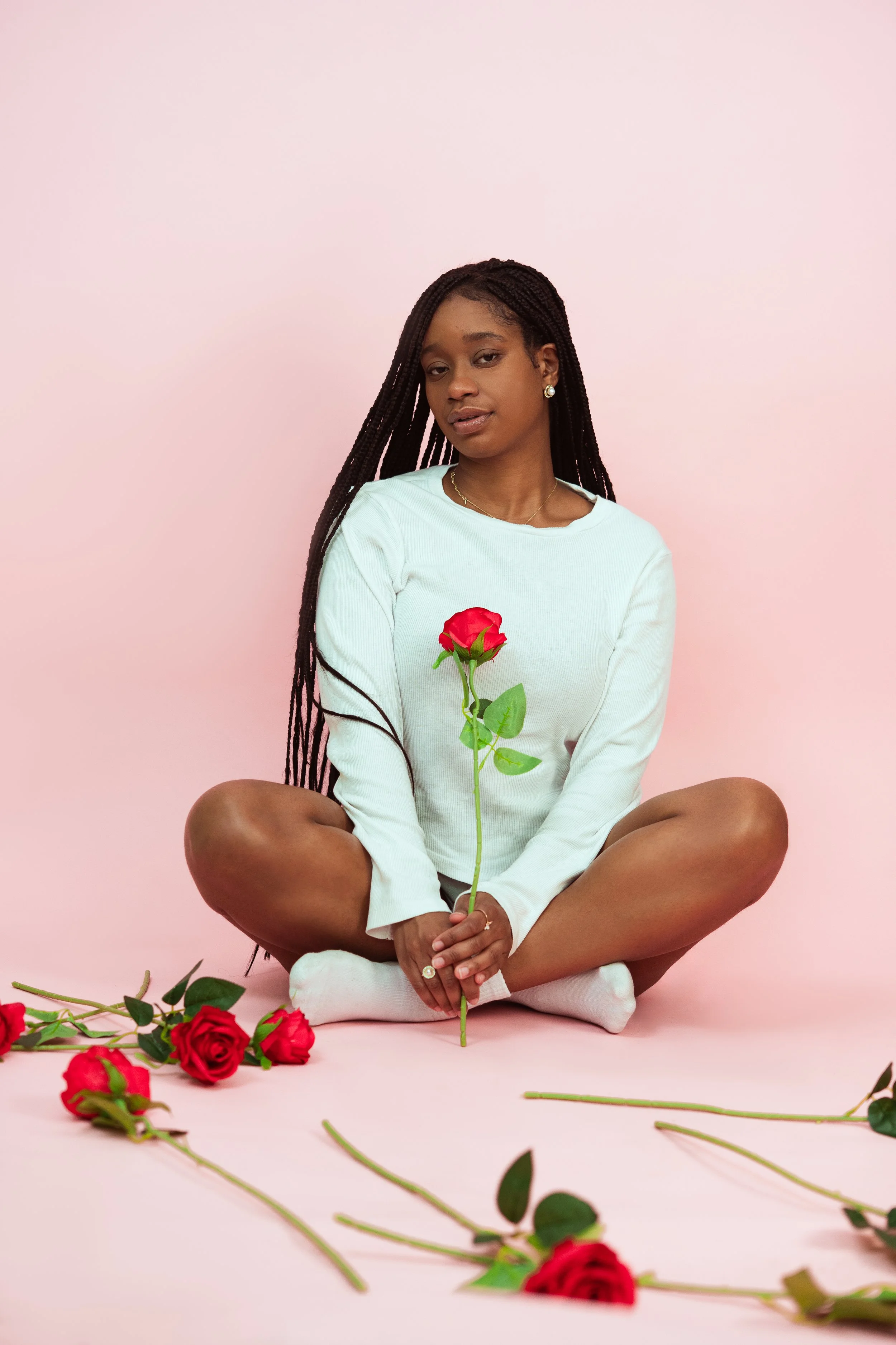 A woman sitting cross-legged on a pink background, holding a single red rose. Several other red roses are scattered around her on the floor.