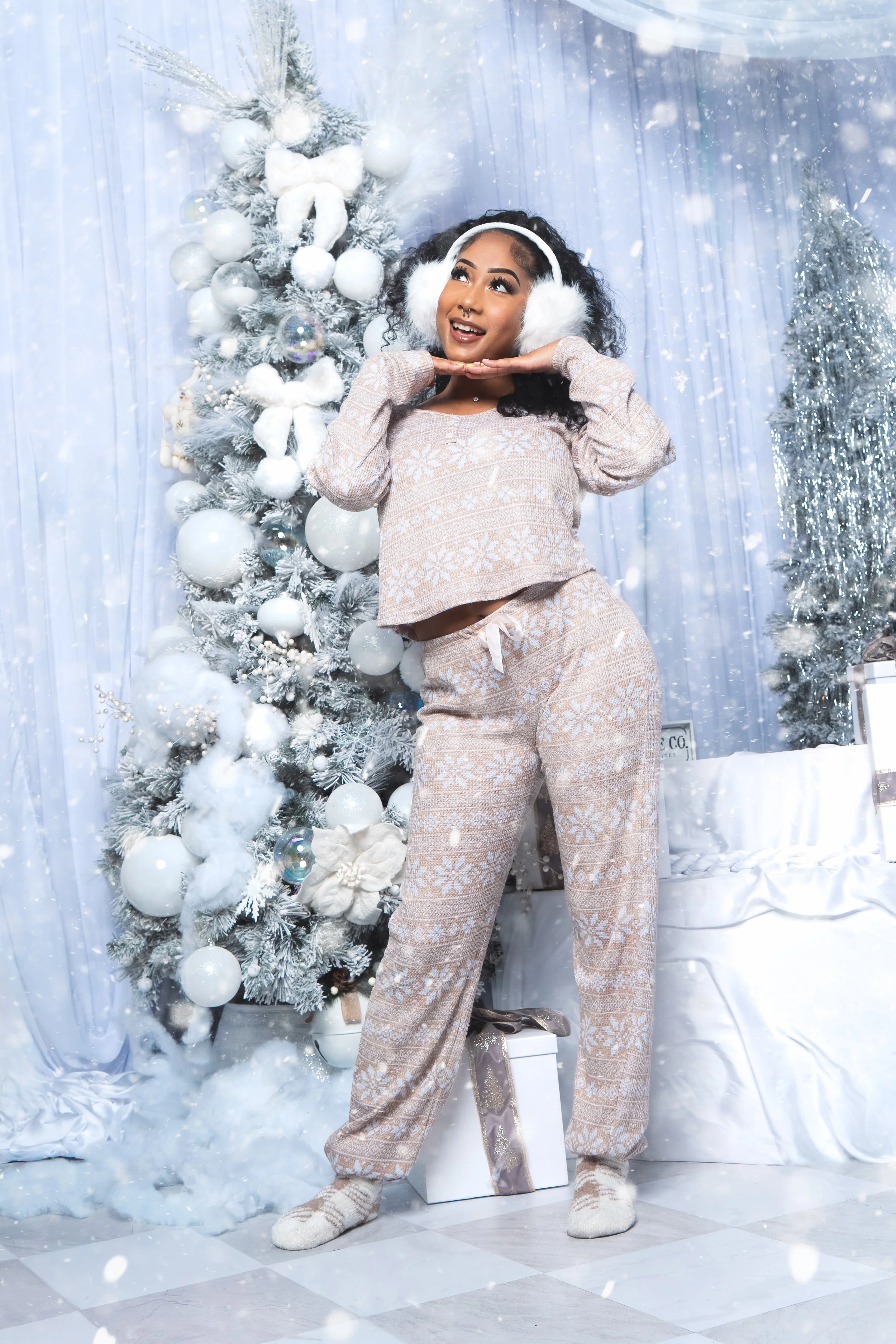 A woman in beige holiday pajamas and earmuffs standing in front of a decorated white Christmas tree, with snow and wrapped gifts around her.