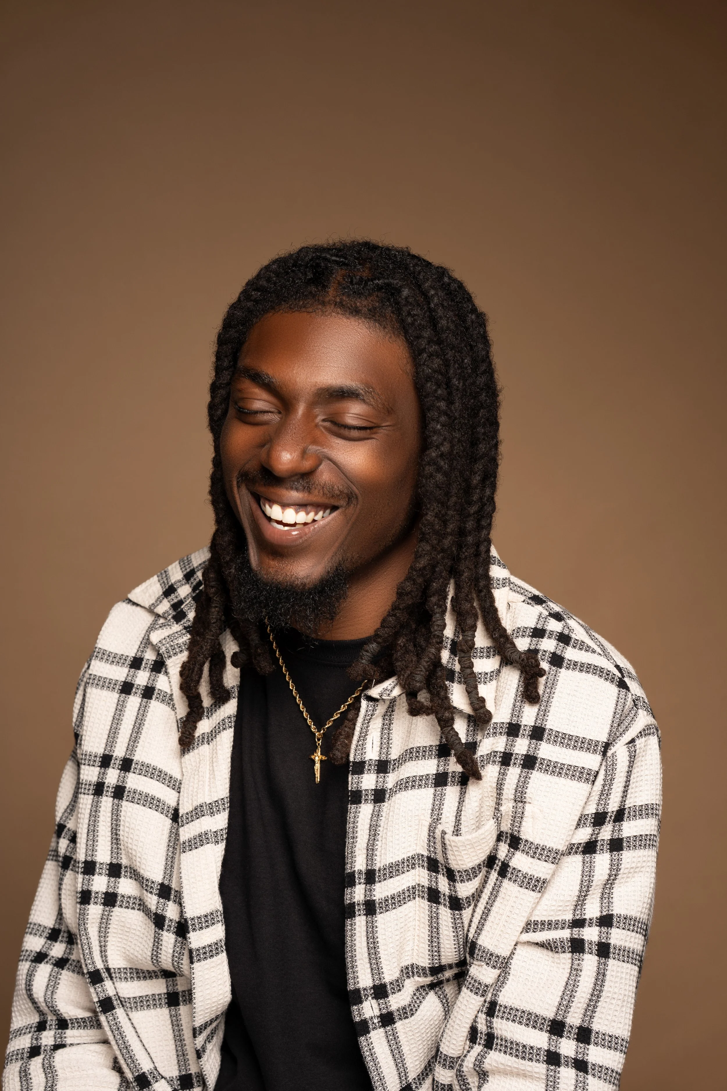 A man with long dreadlocks smiling and looking down, wearing a black shirt, a plaid jacket, and a gold cross necklace, against a brown background.