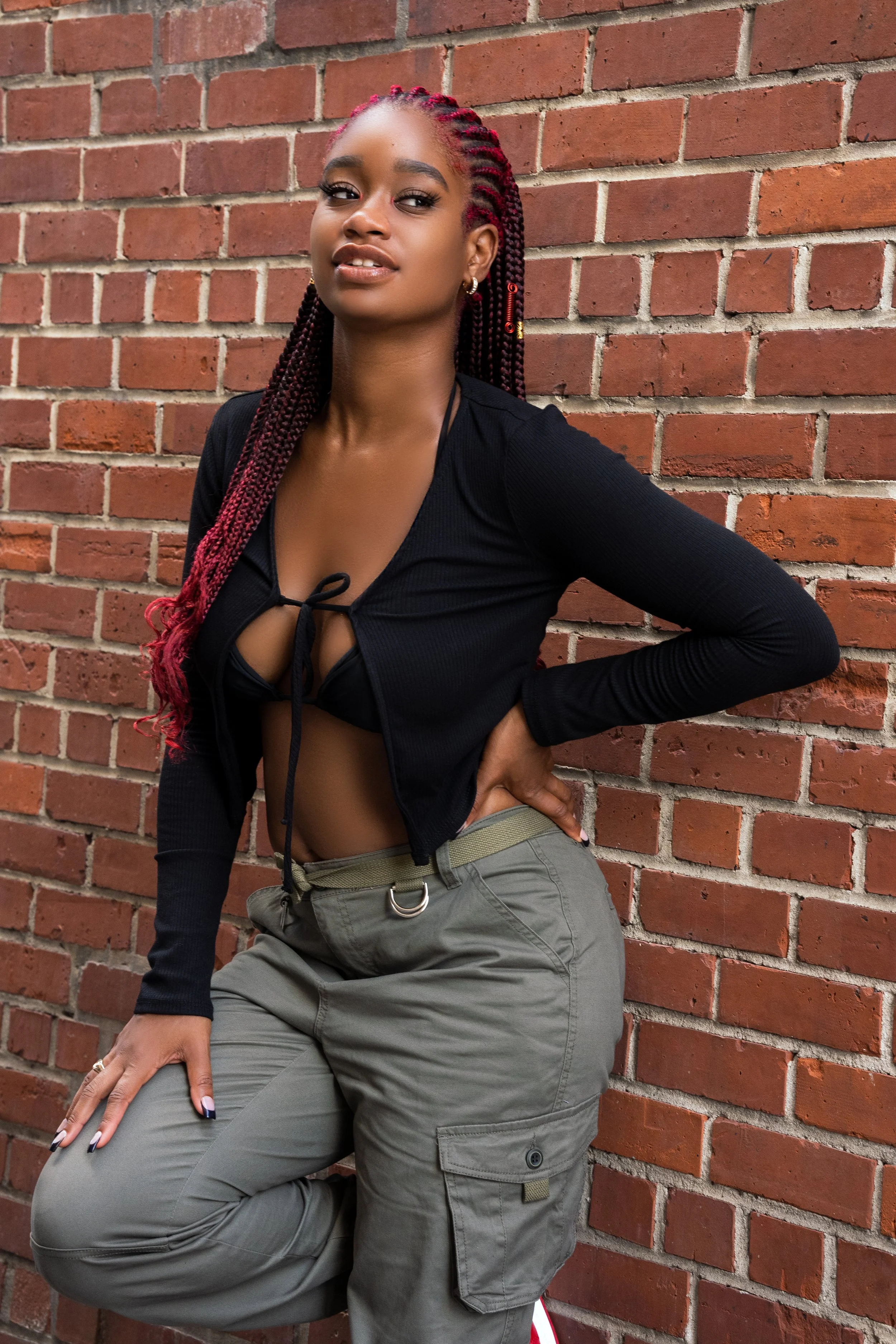 Young woman with red braided hair posing against a brick wall, wearing a black crop top and olive green cargo pants.