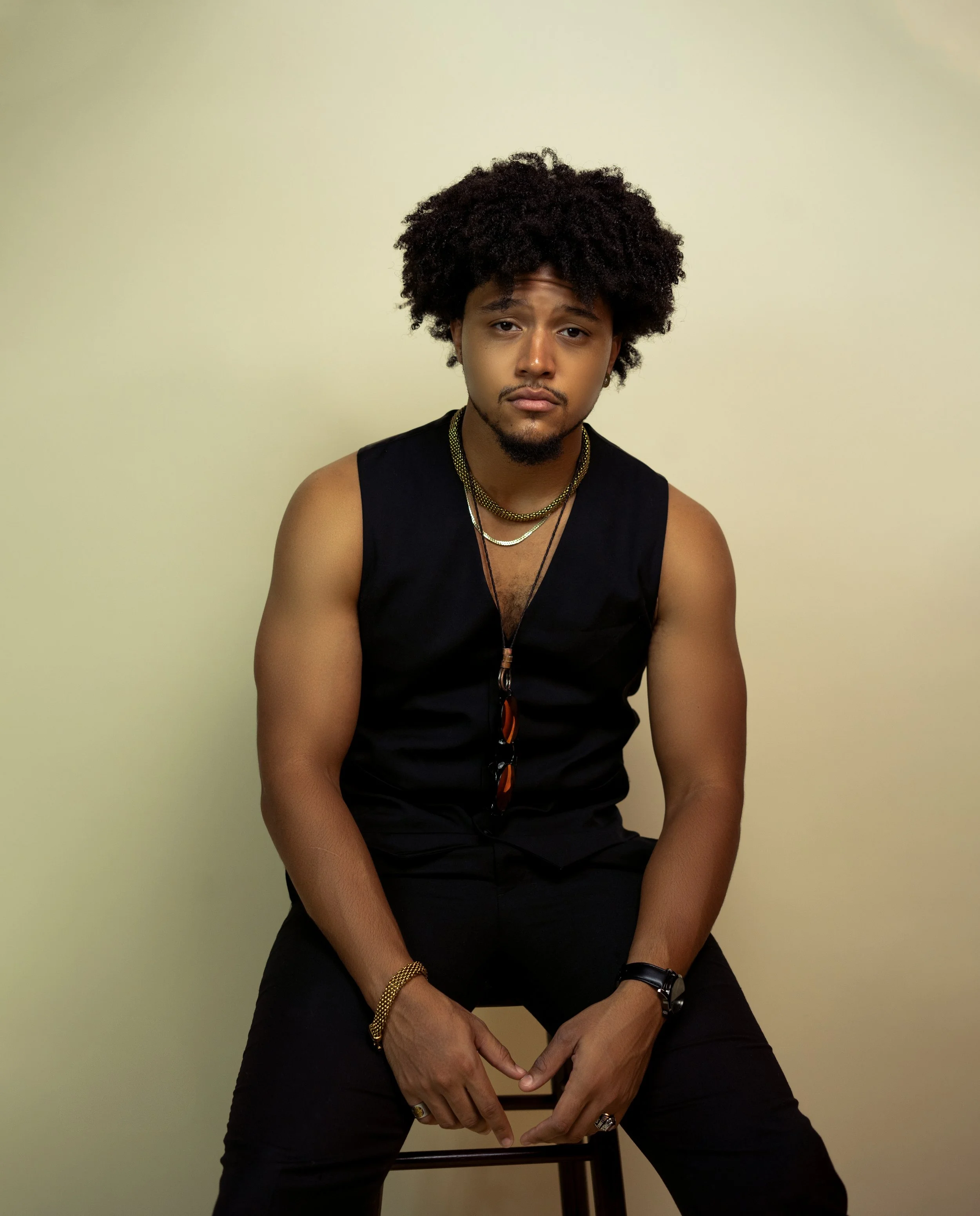Young man with curly hair sitting on a stool wearing black sleeveless top, gold jewelry, and sunglasses hanging from shirt, against a plain beige background.