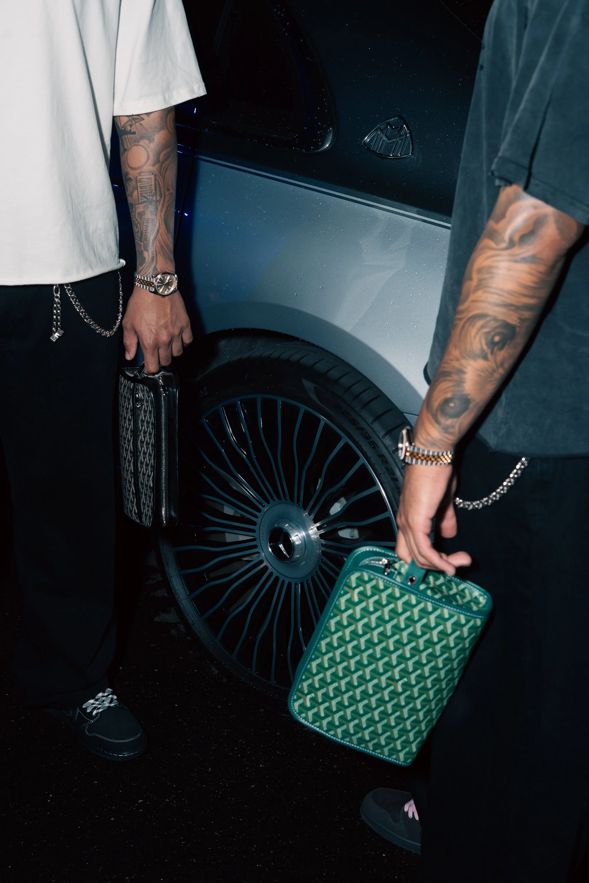Two men with tattoos holding patterned bags, standing next to a luxury black car with alloy wheels.
