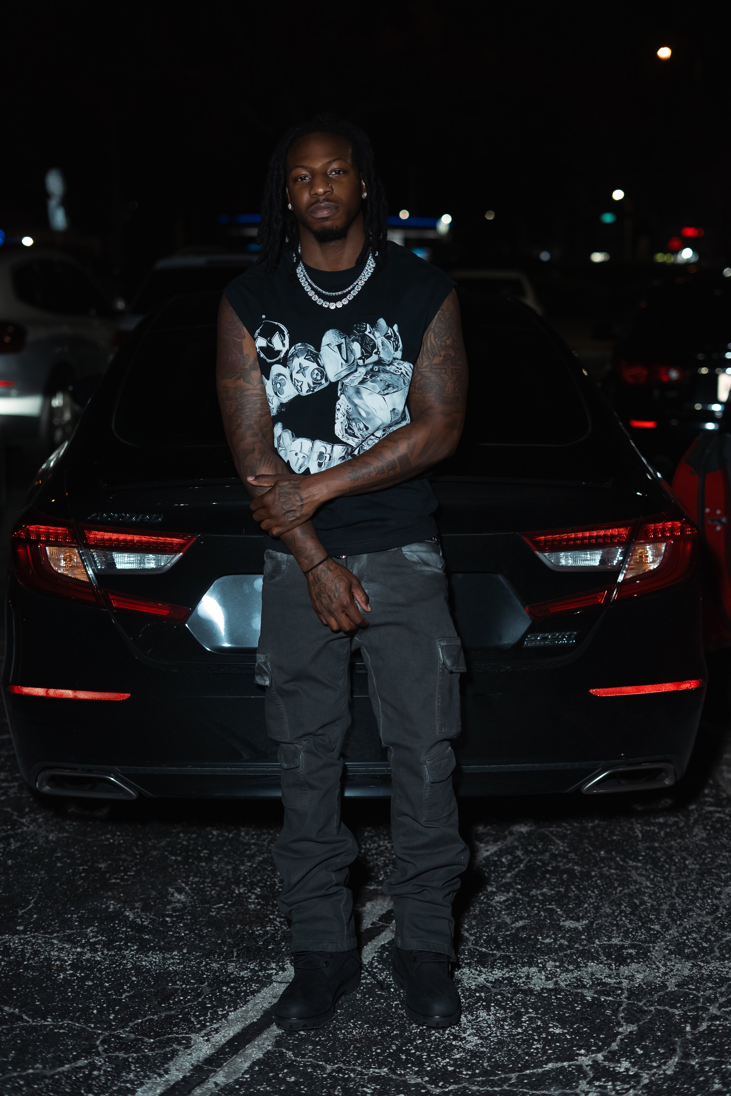A young man with long dreadlocks, tattoos, and earrings stands in front of a black car at night, crossing his arms and looking at the camera. He is dressed in a sleeveless graphic shirt, cargo pants, and black boots. The background shows a parking lo