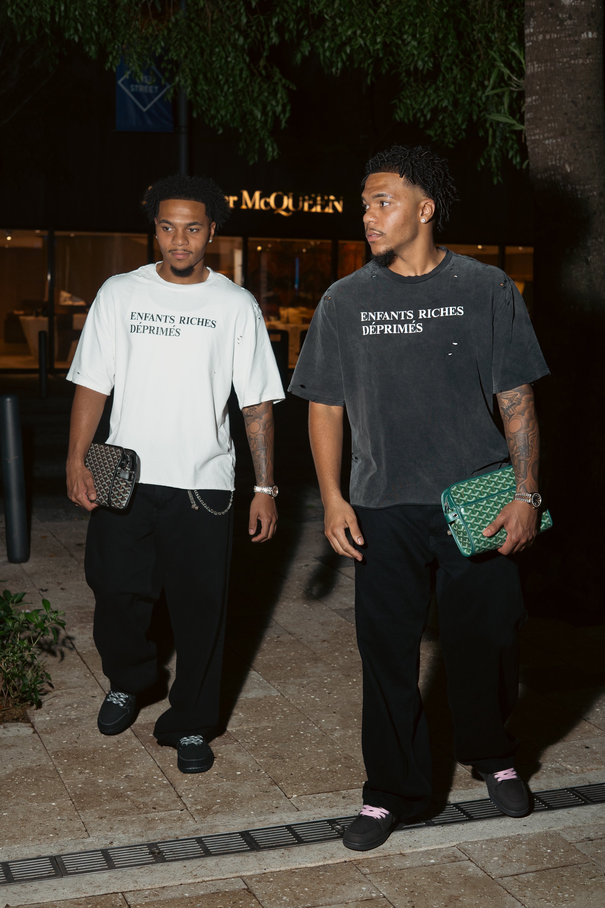 Two young men walking outside at night, wearing distressed T-shirts with French text, carrying designer clutches, and dressed in black pants and sneakers.