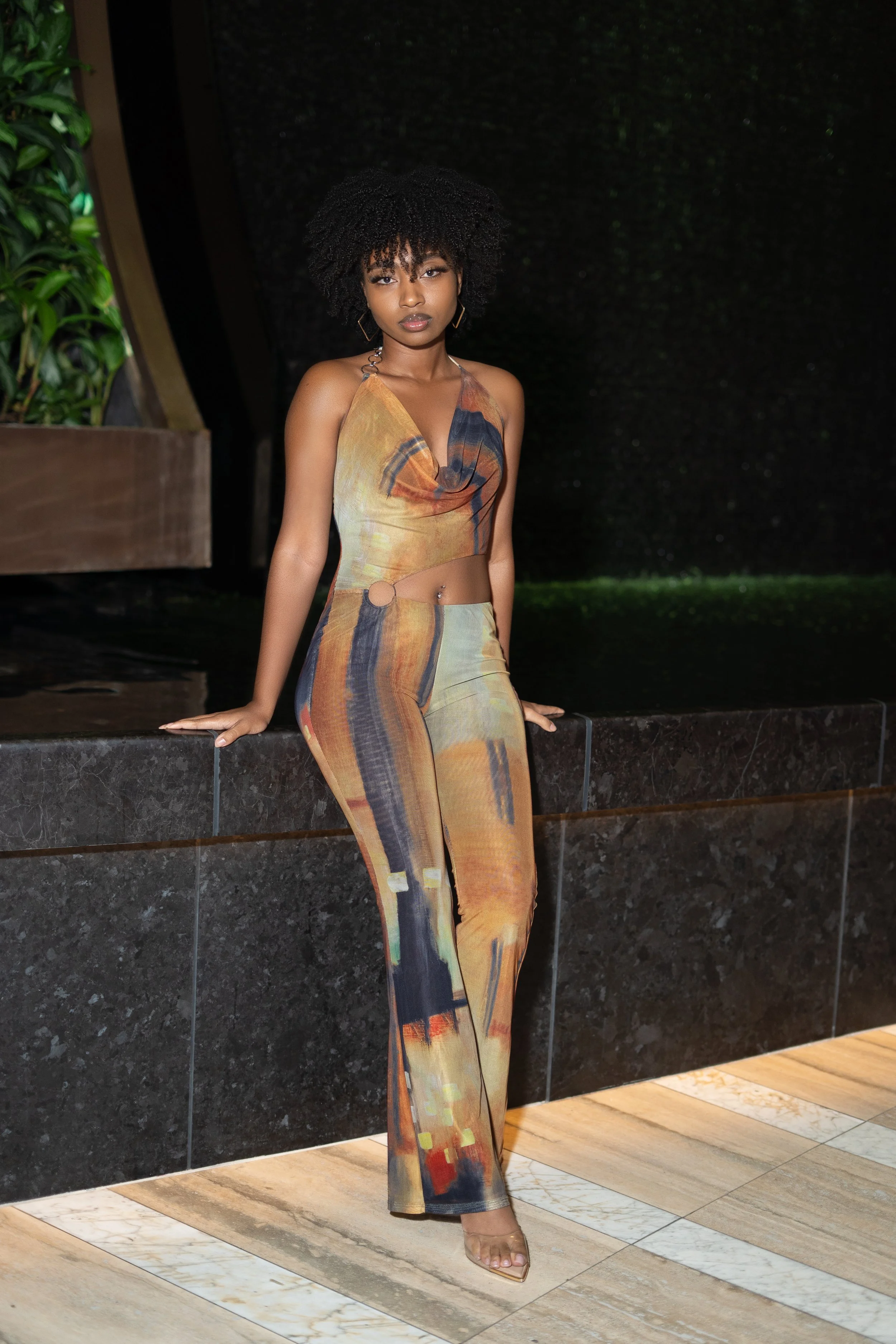 A woman in a colorful, abstract-patterned two-piece outfit with a crop top and high-waisted pants, standing indoors against a dark background, leaning on a black marble ledge.