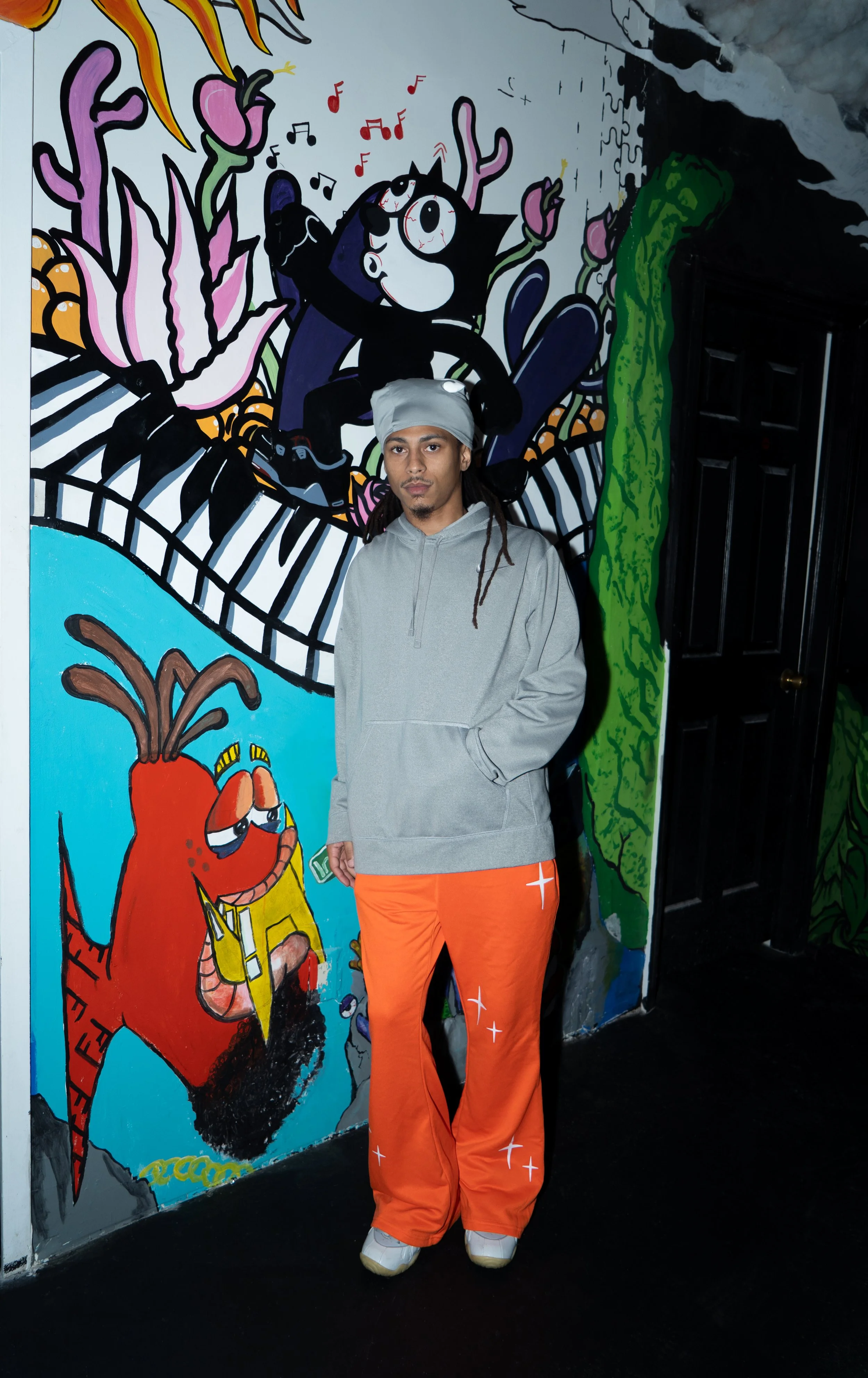 A man with long dreadlocks wearing a gray hoodie, gray headscarf, and orange pants with star patterns standing in front of a colorful street art mural featuring cartoon characters and vibrant plants.