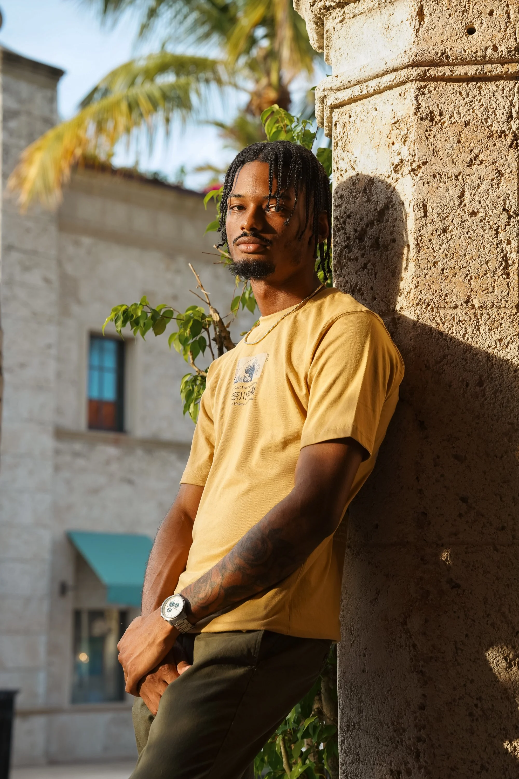 A young man with dreadlocks, wearing a yellow T-shirt, standing outside against a sunlit stone wall, with palm trees and a building in the background.