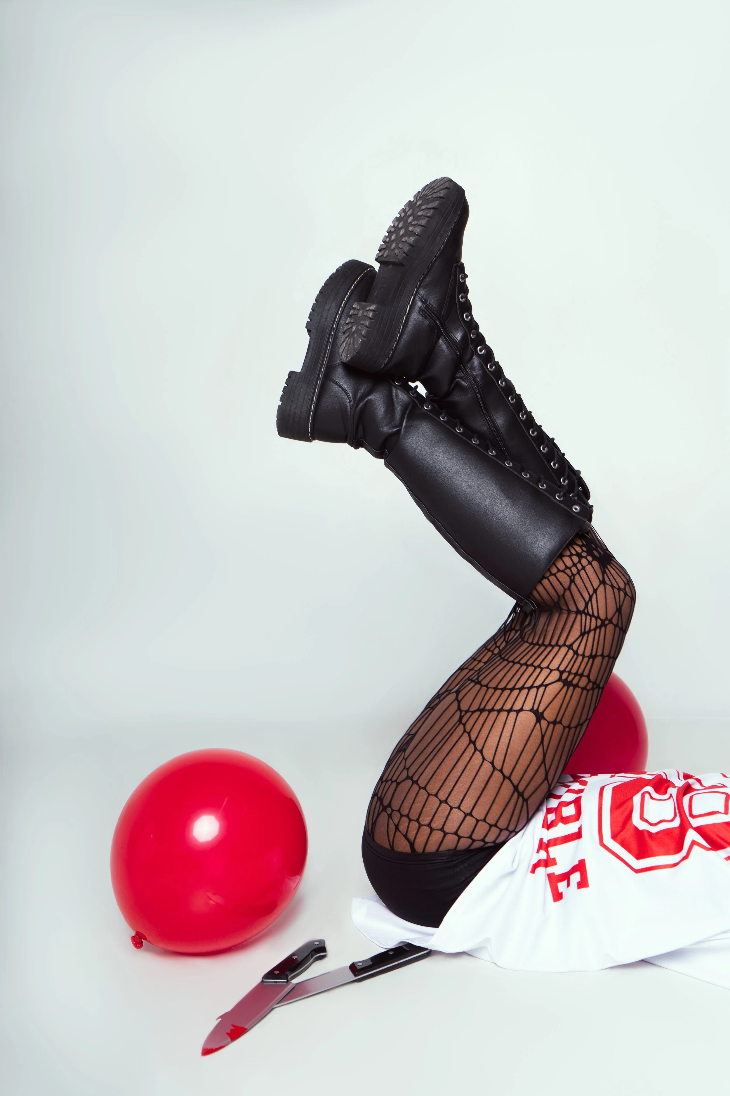 Person lying on the floor with legs raised, wearing fishnet stockings, black combat boots, and a sports jersey, with a red balloon and a folding knife nearby.