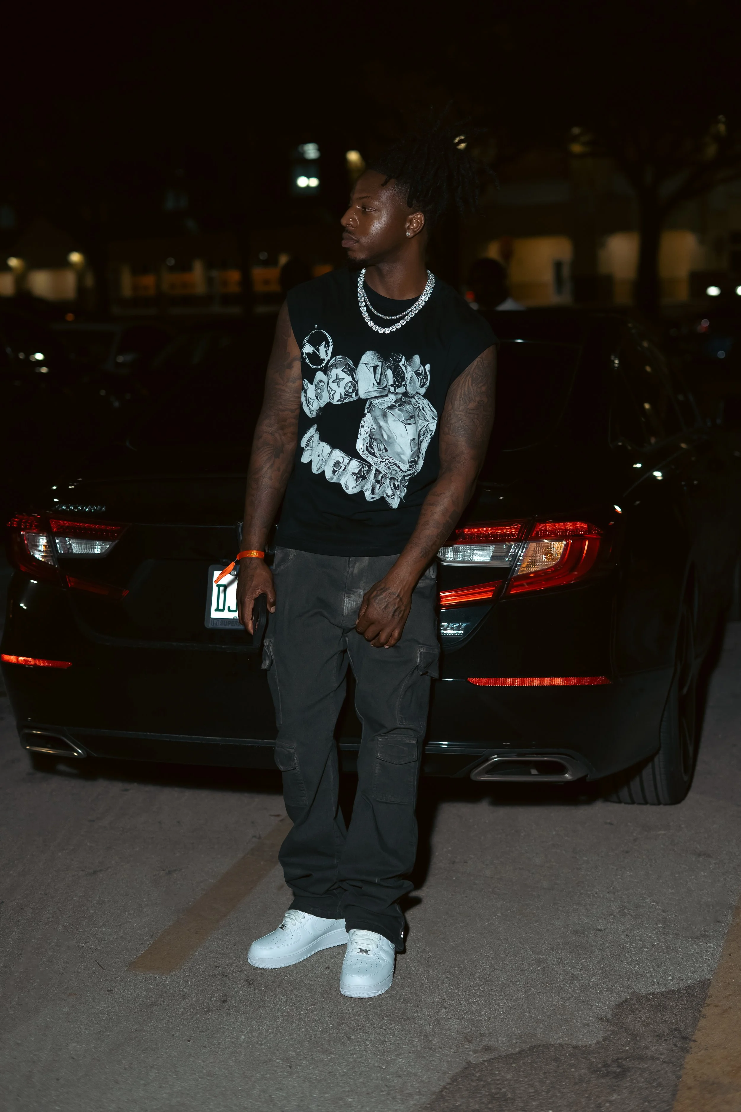 A young man with dreadlocks and tattoos stands in front of a black car at night, wearing a black sleeveless shirt, cargo pants, and white sneakers.