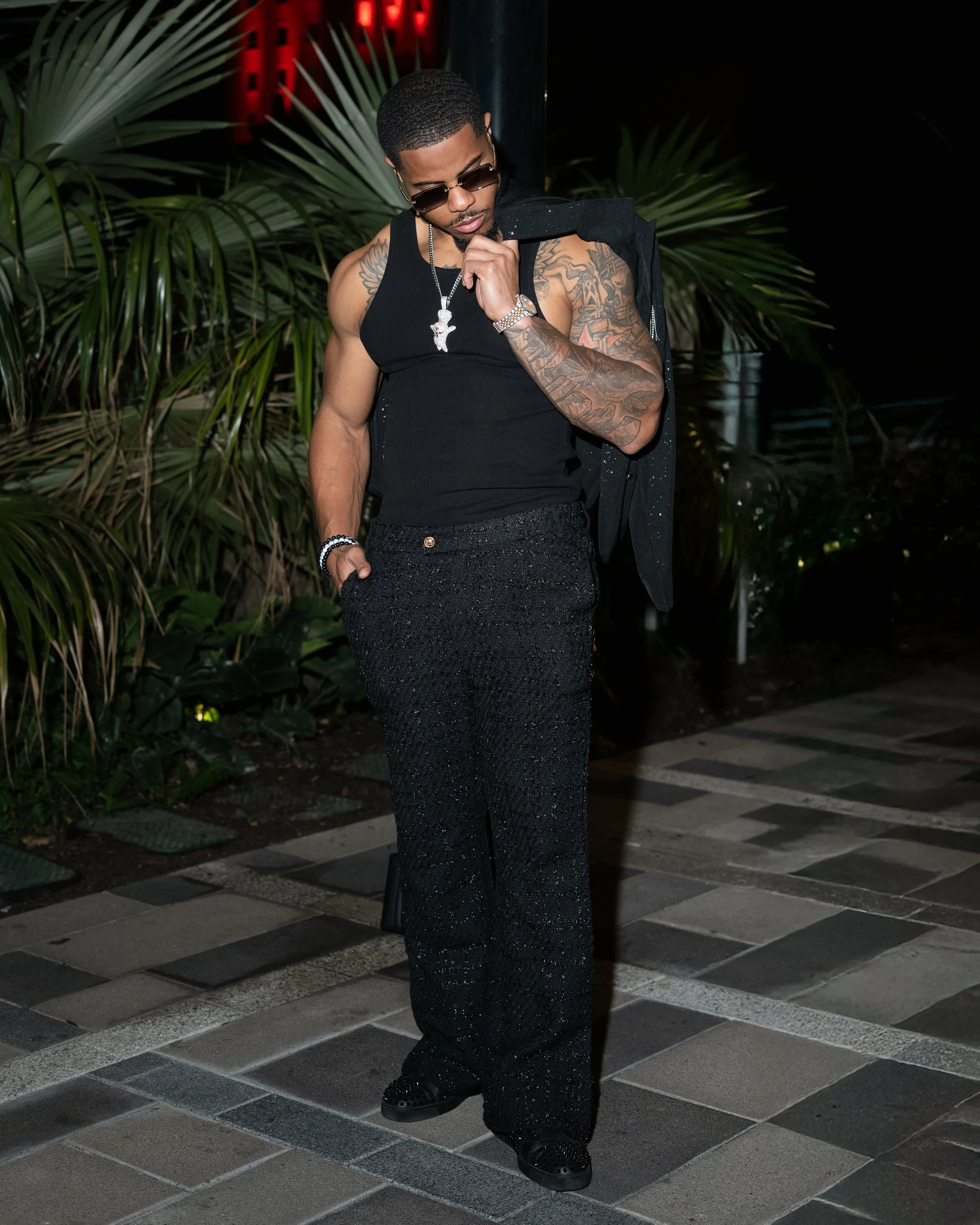 A man in black sunglasses, a black tank top, textured black pants, and black shoes standing outdoors at night, with palm plants and a dark background behind him.