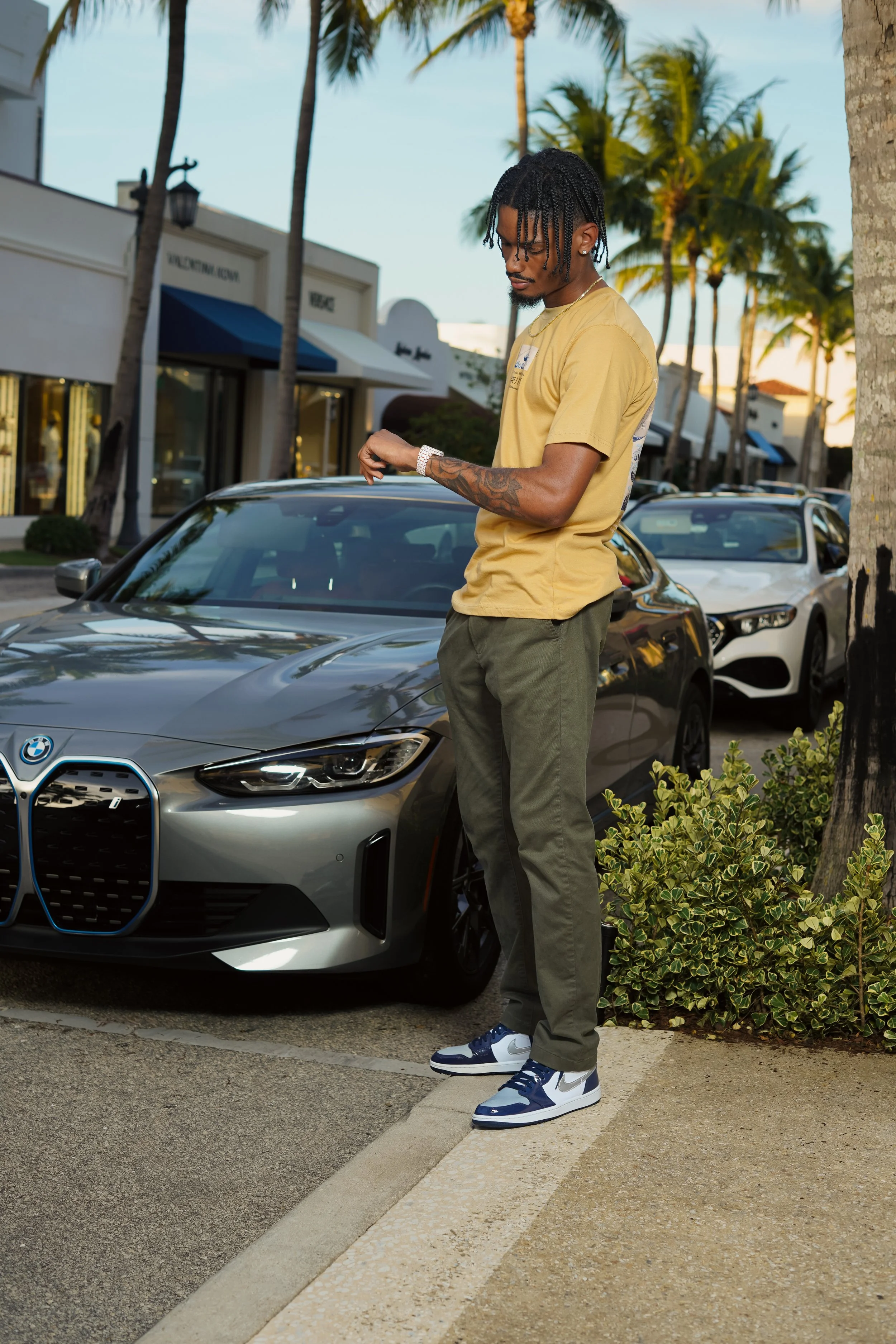 A young man with dreadlocks, wearing a yellow t-shirt, green pants, and Nike sneakers, stands next to a gray BMW parked on the street with palm trees and storefronts in the background during the evening.