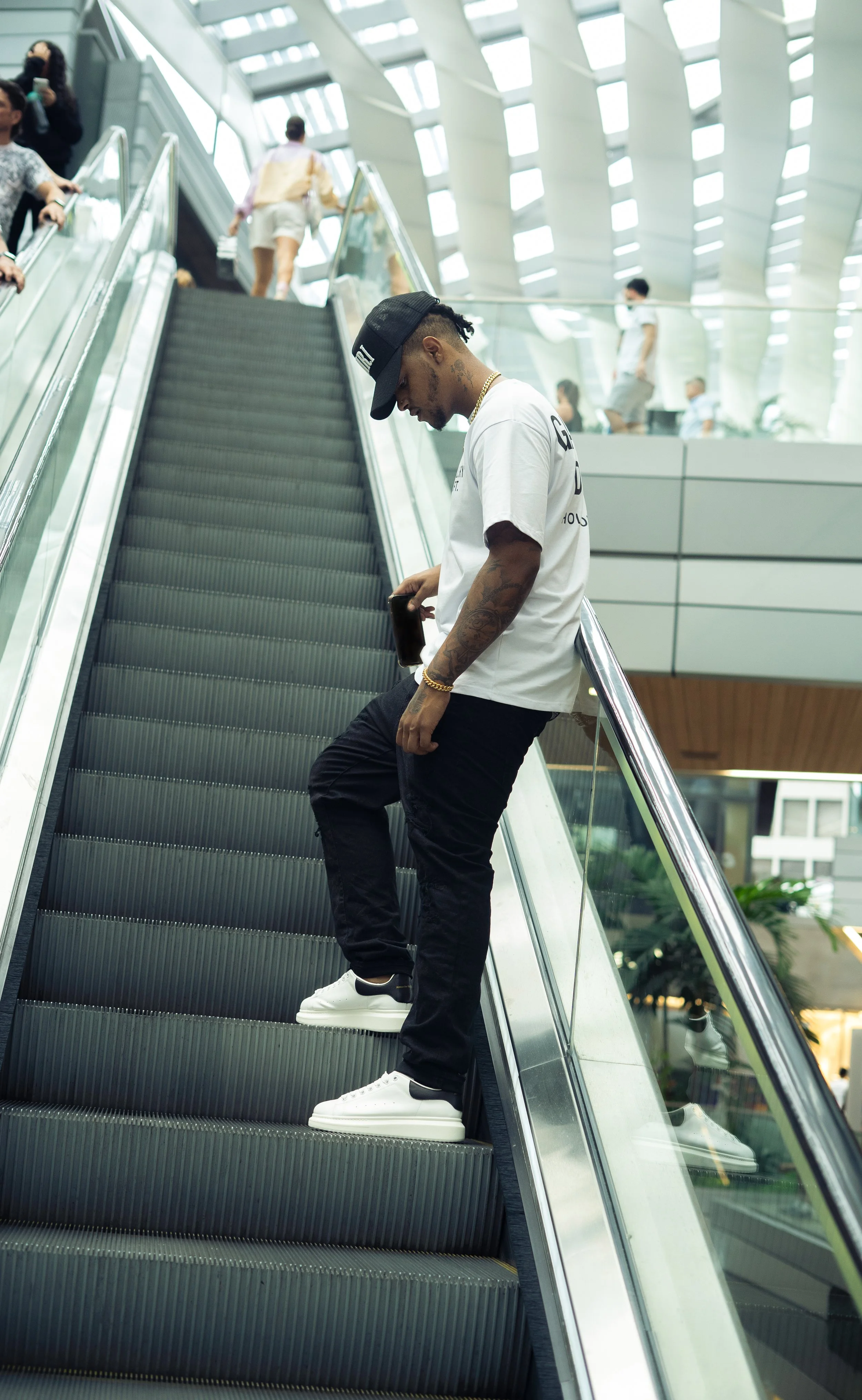 Young man with tattoos, wearing white t-shirt, black pants, white sneakers, a black cap, and gold jewelry, standing on an escalator in a modern indoor shopping mall or airport.