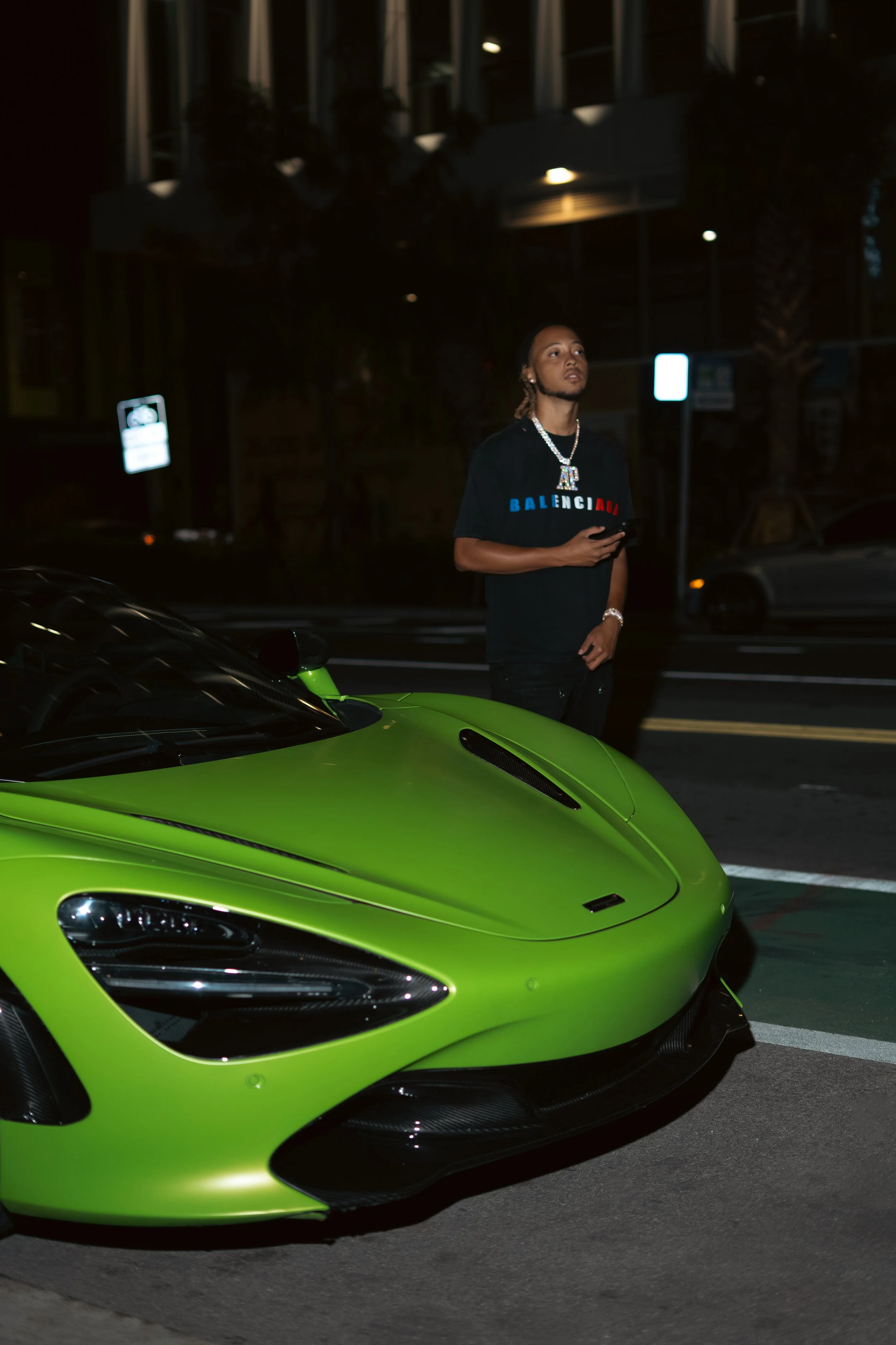 A young man stands beside a bright green luxury sports car on a city street at night, holding a phone and looking upward.