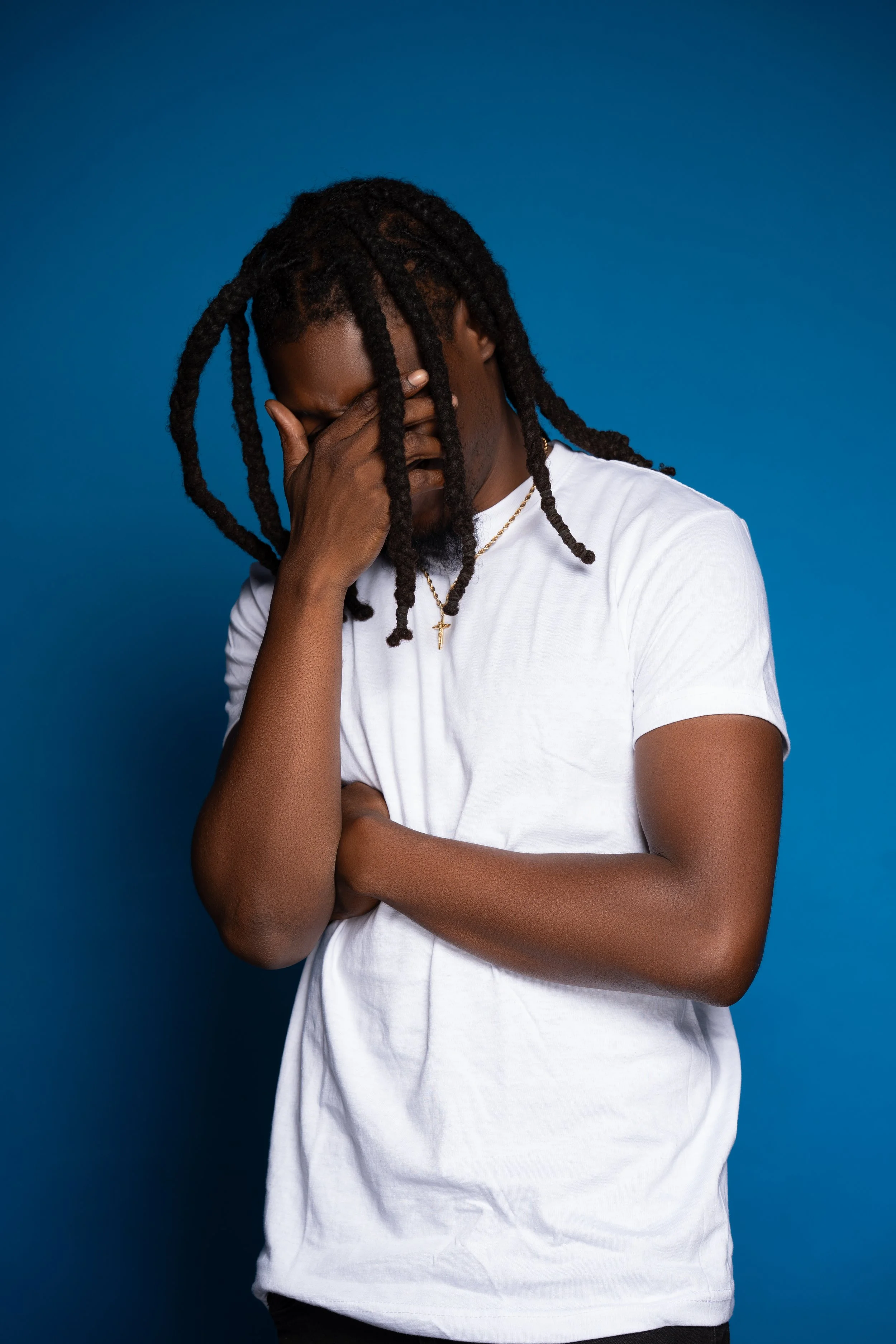 A man with dreadlocks wearing a white t-shirt, with his hand covering his face, standing against a blue background.