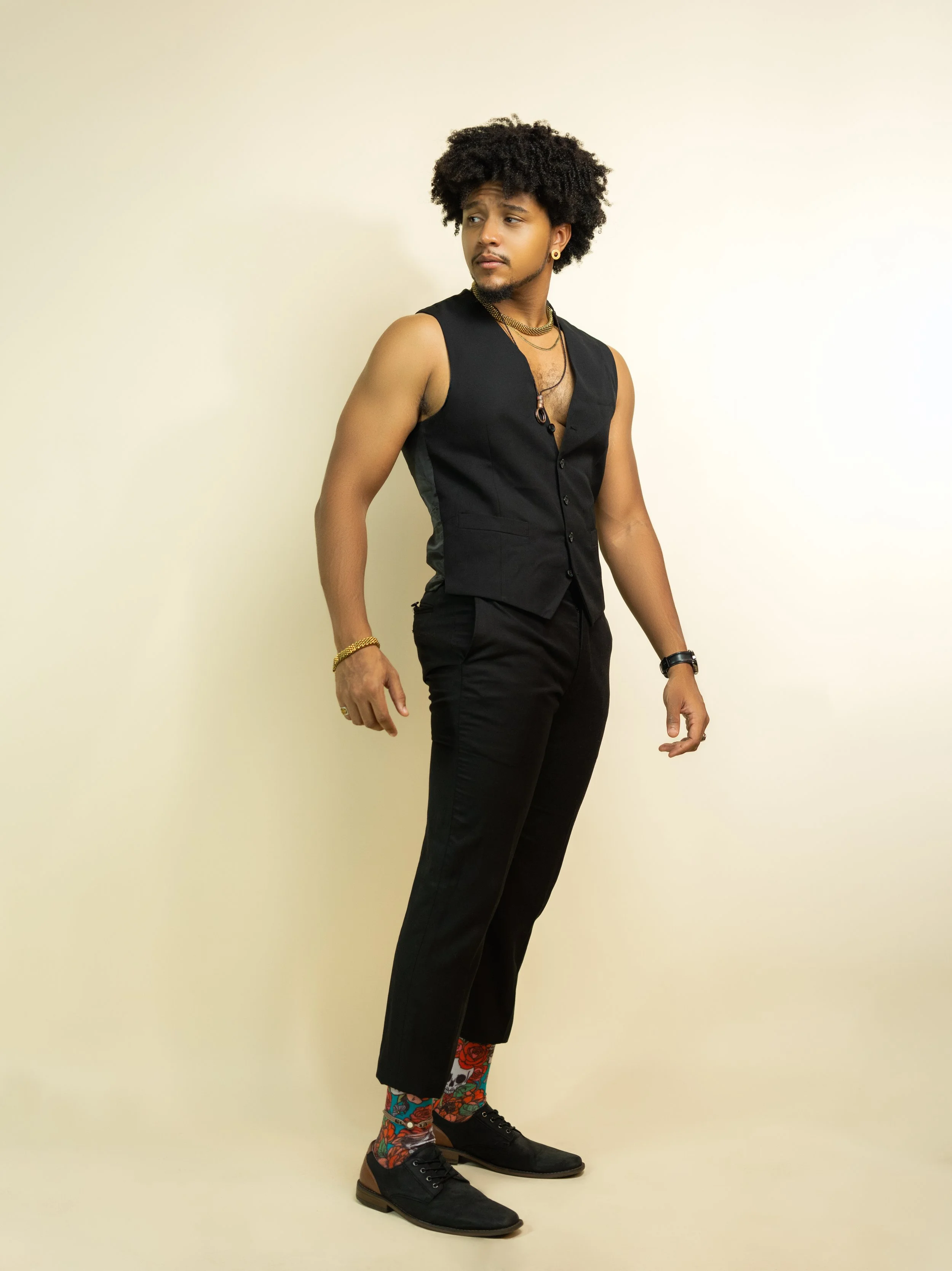 A young man with curly hair wearing a sleeveless black vest, black pants, black shoes, and colorful floral socks, standing against a plain beige background.