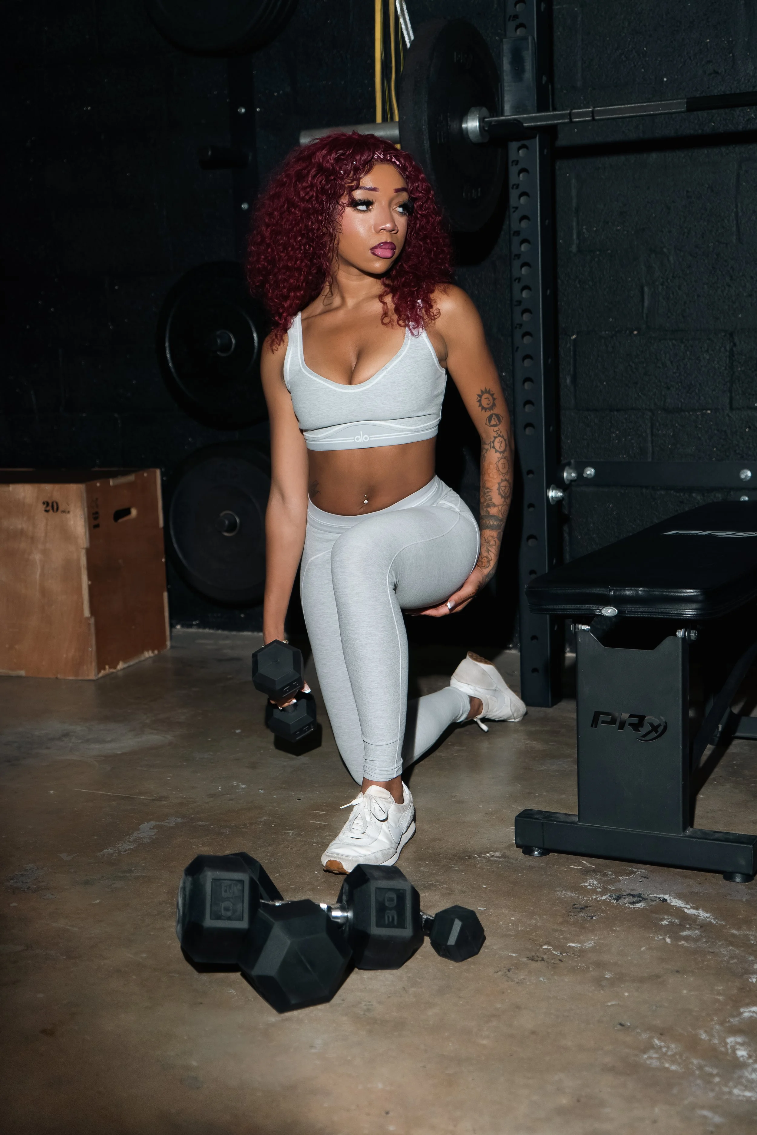 A young woman with curly red hair in workout attire kneeling on one knee in a gym, holding a dumbbell in her right hand, with weight plates and gym equipment in the background.