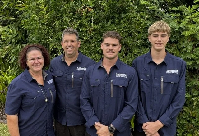 Four people wearing navy blue shirts with 'Authentic' embroidered on the chest, standing outdoors in front of lush green foliage, smiling for the photo.