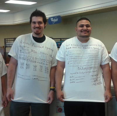 Two young men wearing white t-shirts with handwritten notes stand in front of a beige wall with a sign that reads 'The Wheel'.