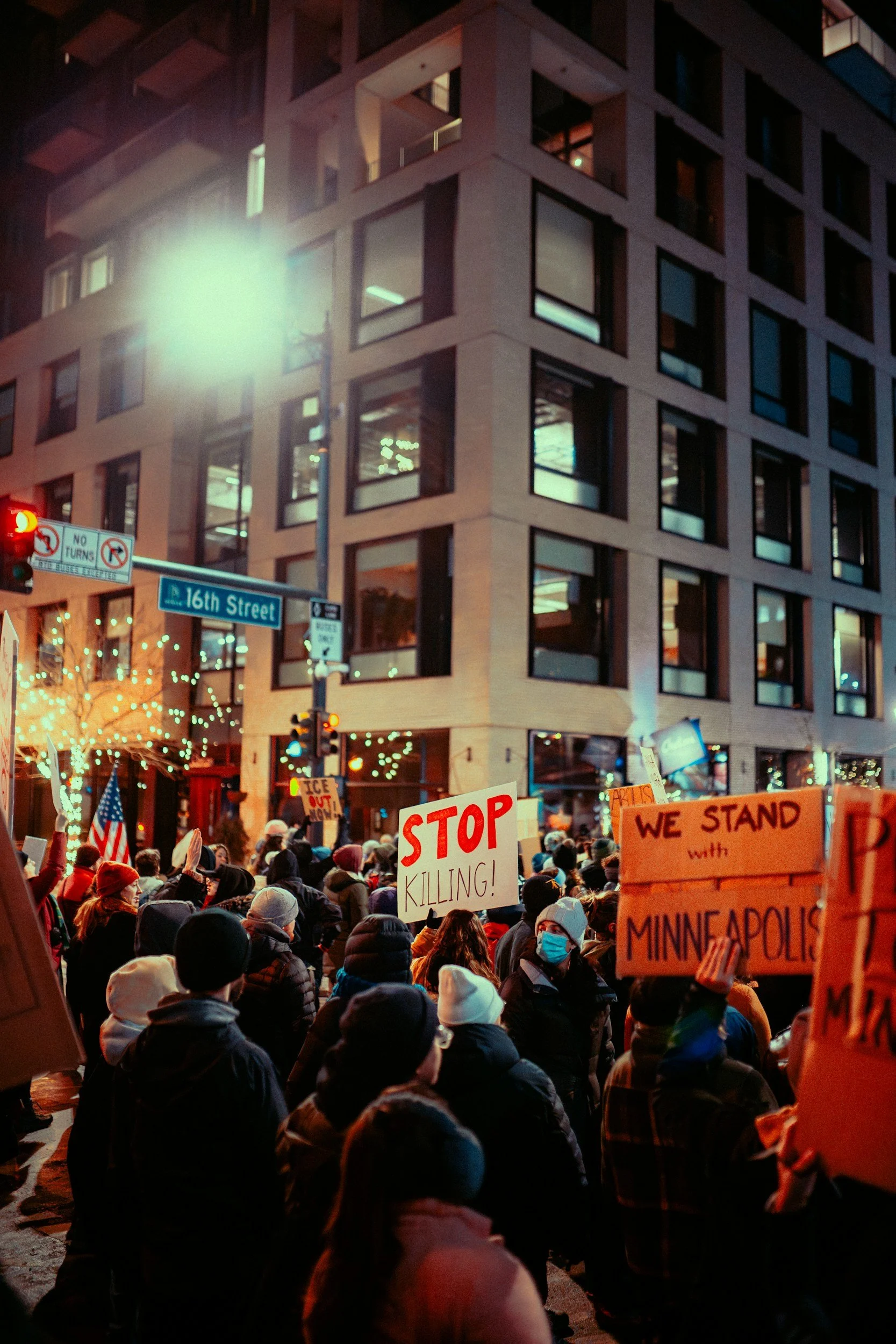 Protesters gathered on a city street holding signs that say "STOP KILLING!" and "WE STAND with MINNEAPOLIS." The scene is at night with building lights and decorative streetlights.