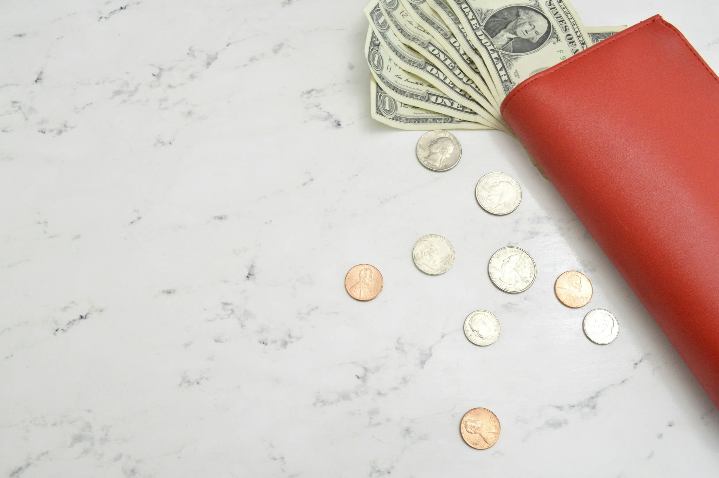 A red wallet tipped over on a white marble surface, with US dollar bills and coins spilling out.