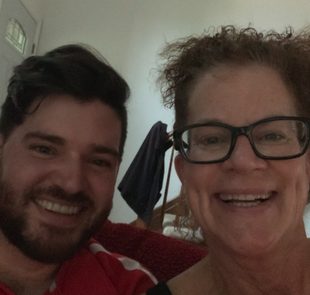 A smiling woman with curly hair and glasses taking a selfie with a smiling man with dark hair and beard.