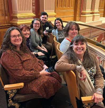 Six people sitting in a row of seats in a theater or auditorium with ornate woodwork and railing behind them, smiling at the camera.