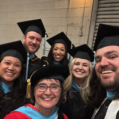 Six graduates in caps and gowns, smiling for a group photo at a graduation ceremony.