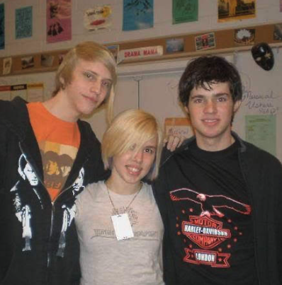 Three teenagers standing together inside a classroom with colorful posters and decorations on the wall behind them.