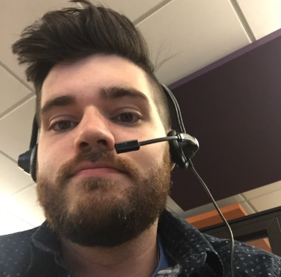 Close-up of a man with dark hair, a beard, wearing a headset, in an office setting.
