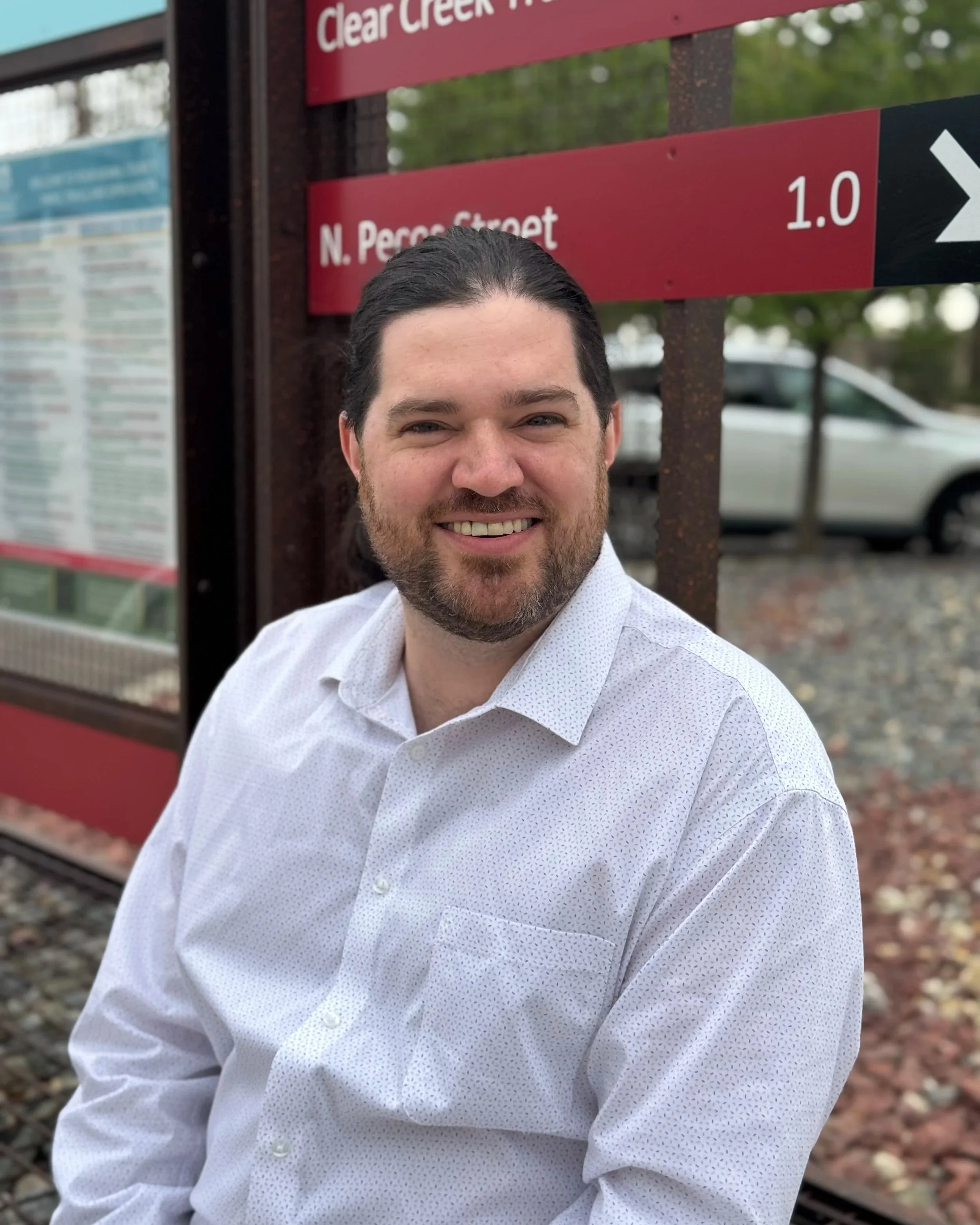 Smiling man in a white shirt sitting outdoors near a red sign with city street signs and vehicles in the background.