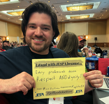 A smiling man holding a yellow sign at a conference. The sign reads: '1stand with JESPA because... Every professional deserves Respect AND dignity!!!' and includes the name Alex Rykhmann, organization Apple to EA, and the hashtag #jeffcodobetter. The background shows other attendees and conference tables in a large, well-lit room.